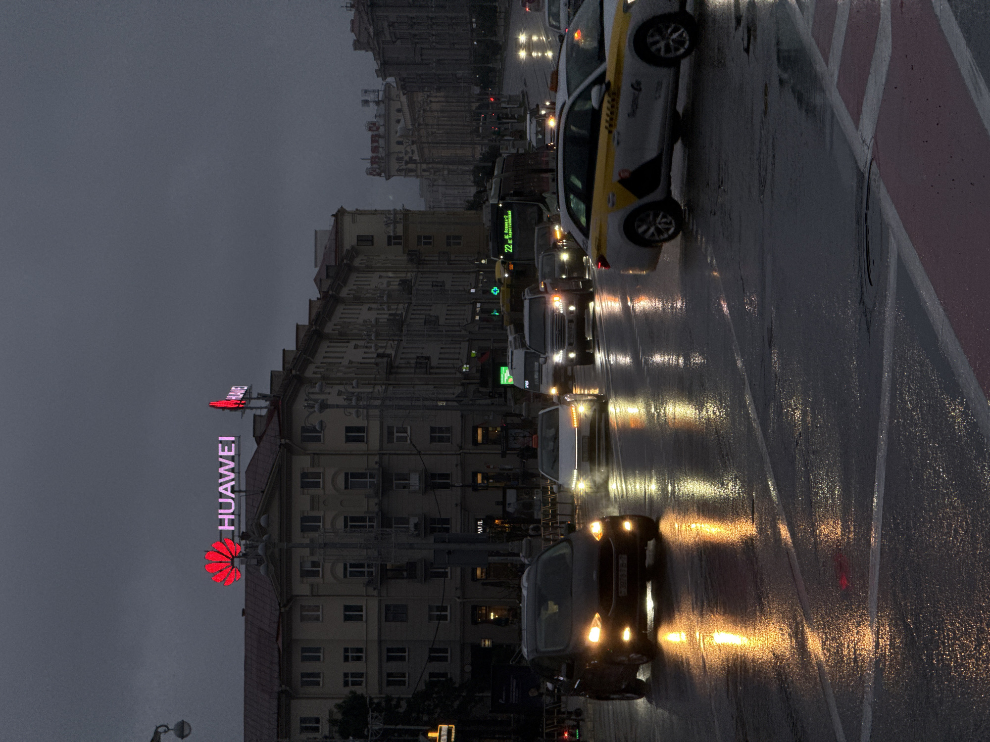 The image captures a street scene at dusk or night in Minsk, Belarus, characterized by wet asphalt reflecting the lights of vehicles and buildings. The sky is overcast and dark.
Prominently featured on the facade of a large, multi-story building is a bright pink neon sign that reads "HUAWEI" with the distinctive red Huawei logo preceding it. Another similar, smaller Huawei sign is visible further down the building. The building itself is a light-colored, classical architectural style with numerous windows and cornices.
On the street, several vehicles are present. In the foreground left, a dark-colored car with its headlights on is driving. To its right, a white car with its headlights also illuminated is proceeding. Further down the street, a yellow and white taxi is visible, as is a bus with a lighted route display showing "22 БОЛЬШ. КОЛЬЦА - Хрустальная". Other cars and a large truck are also visible in the middle ground and background.
The wet road surface creates strong reflections of headlights, taillights, and building lights, adding to the atmospheric, dimly lit feel of the scene. Red and white markings are visible on the road surface in the foreground, suggesting a crosswalk or intersection. Streetlights and traffic lights are also visible, contributing to the overall illumination. The environment appears to be a well-developed urban area with substantial architecture and active traffic, likely a main thoroughfare in the city.