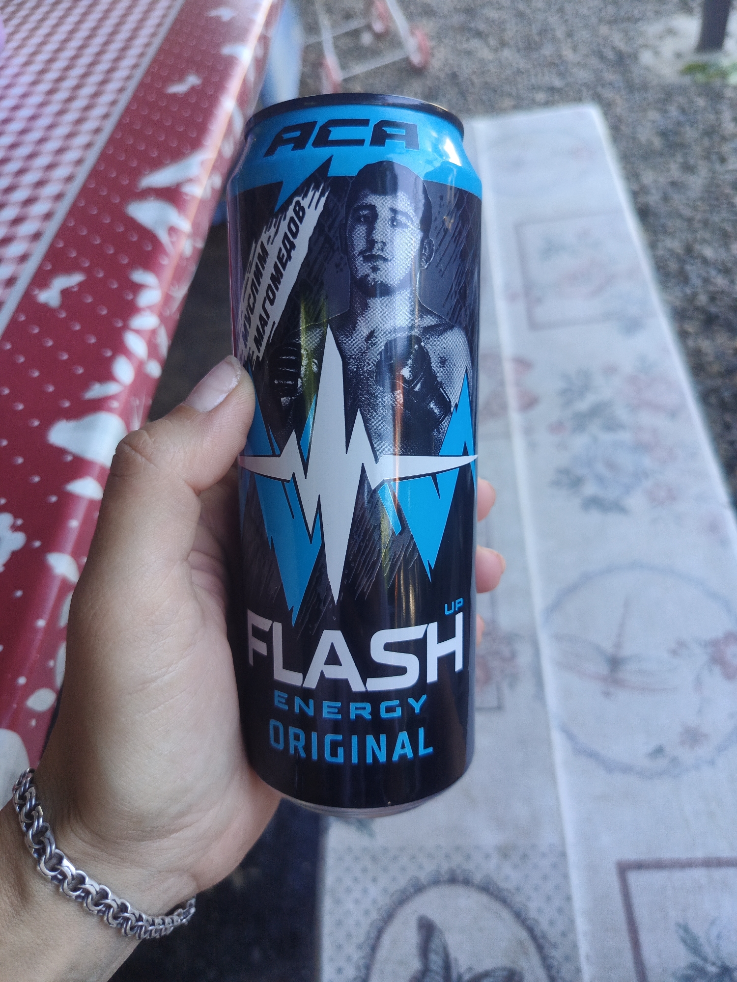 A hand holds a can of "FLASH ENERGY ORIGINAL" drink. The can has a black background with blue and white graphics, including a lightning bolt design and a portrait of a man. The can is positioned in front of a red and white checkered surface and a blurred background. The scene appears to be outdoors, possibly on a table. There is no discernible information within the image to link it to the city of Staronizhestebliyevskaya, Russia.