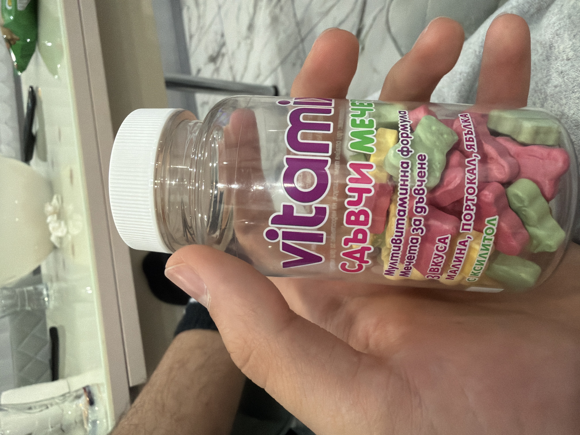 A close-up shot shows a clear plastic bottle of gummy vitamins held by a person's right hand. The bottle is filled with colorful, bear-shaped gummies in shades of pink, green, and yellow. The label on the bottle features the word "vitami" in large purple letters, followed by smaller text in Bulgarian that translates to "gummy bears" and "multivitamin formula for chewing." Beneath that, it lists flavors such as "strawberry, raspberry, orange, apple," and "xylitol." The background is blurred, showing a table with various items, including glasses and what appears to be a green snack bag. The overall scene suggests an indoor setting, possibly a home environment. There is no specific indication of the location in Podgumer, Bulgaria.