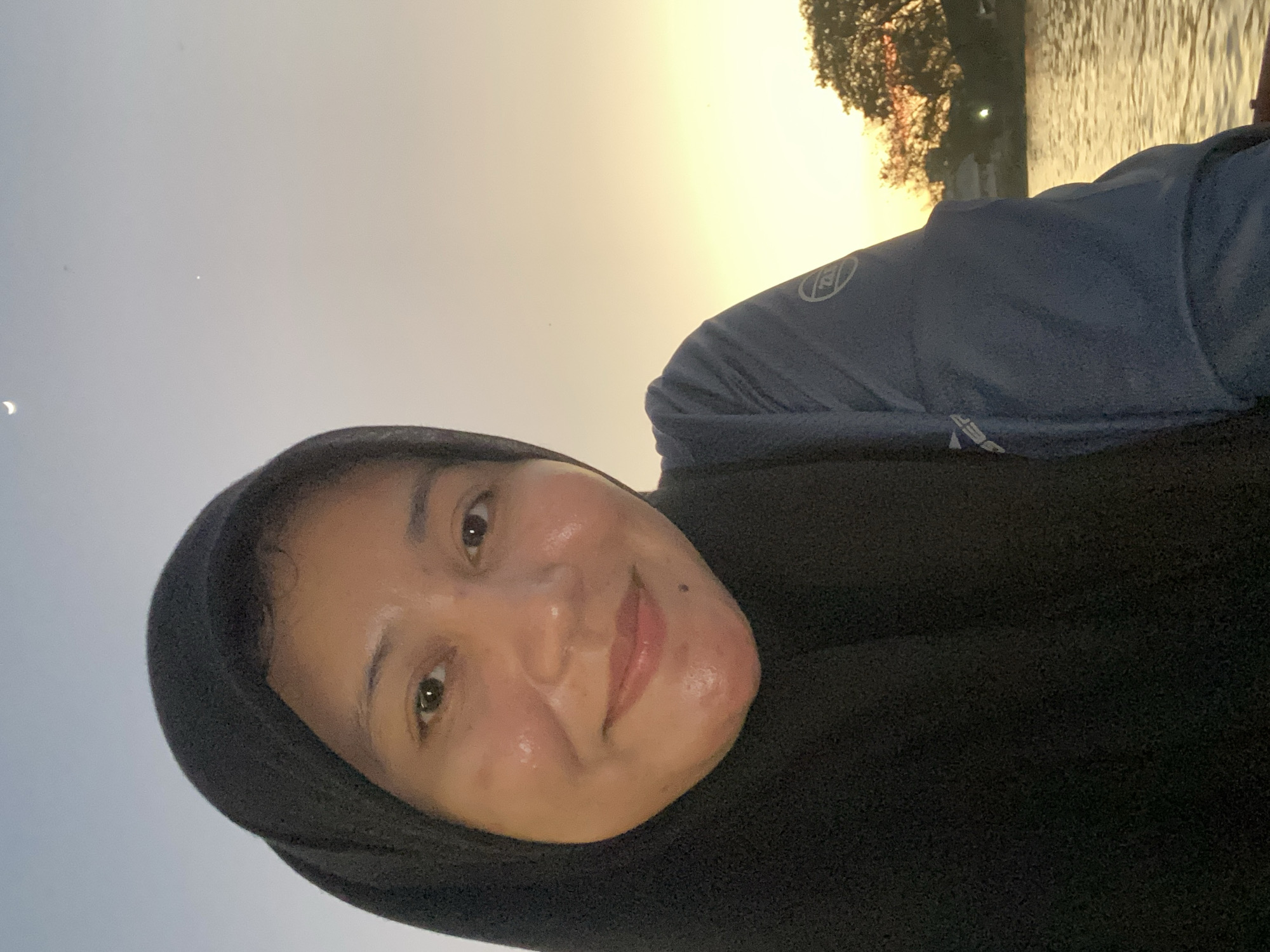A medium shot selfie of a woman wearing a black hijab and a blue long-sleeved shirt is taken outdoors with a body of water and trees in the background. The sky is a gradient of light blue and pale yellow, suggesting either sunrise or sunset. The woman is smiling and looking directly at the camera. The scene is likely taking place in Banjarmasin, Indonesia, known for its extensive river system and lush vegetation. The water appears calm, reflecting the warm tones of the sky. The overall impression is one of peacefulness and natural beauty.