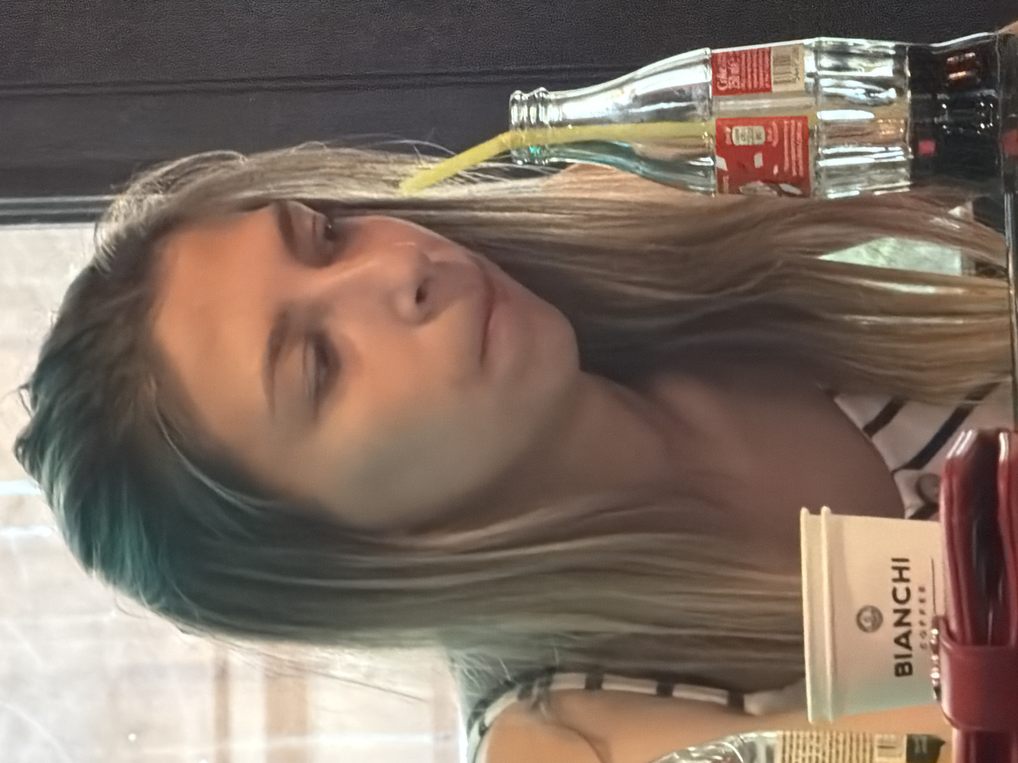 A young woman with blonde hair is sitting at a table in what appears to be a cafe in Gulyantsi, Bulgaria. She is wearing a striped top and is looking down as she drinks from a glass bottle of Coca-Cola with a yellow straw. In front of her is a small white cup with the "Bianchi Coffee" logo, and a red wallet is visible. The background shows a textured dark surface, possibly a wall or furniture, and a window with a blurry reflection. The lighting suggests it is daytime.