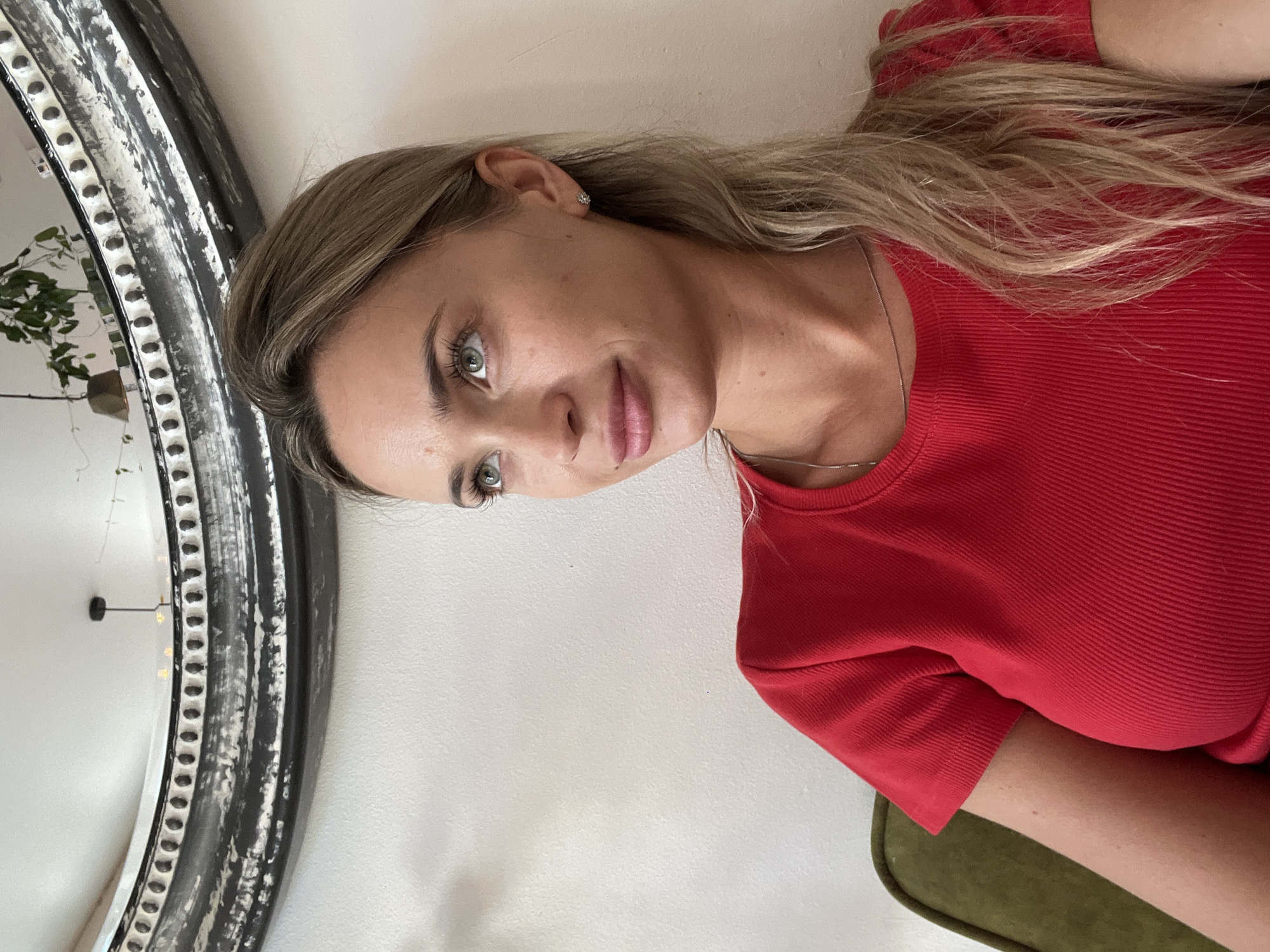 A medium shot shows a blonde woman with green eyes and fair skin, looking up and to her right. She is wearing a bright red, ribbed short-sleeved shirt and a thin silver necklace. Her hair is long and light blonde, pulled back loosely. She has on light makeup, including mascara, eyeliner, and lipstick. A small, sparkling stud earring is visible in her right ear. She is seated on a dark green couch, and the background consists of a white wall with a large, ornate mirror with a distressed black and silver frame. The mirror reflects a ceiling light fixture and some greenery. The scene appears to be indoors, in a residential or commercial space, likely within the city of Odesa, Ukraine.