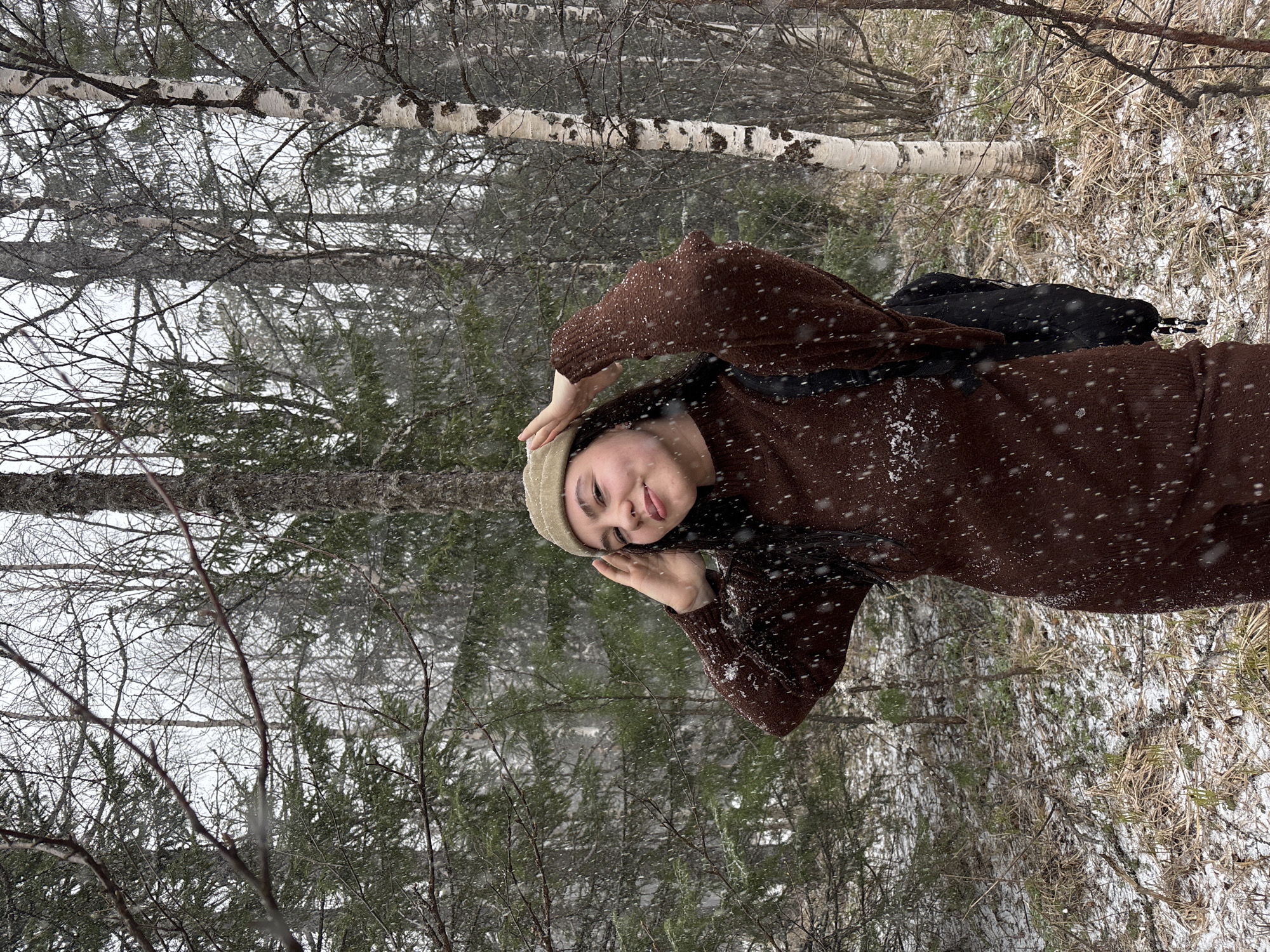 A young woman stands in a snow-covered forest in Kachkanar, Russia, smiling and posing for a photo. She wears a brown sweater dress and a light-colored knit hat, shielding her eyes with her hand. Bare trees and evergreens surround her, with a birch tree visible in the background. Light snow falls. A black bag is slung over her shoulder.
