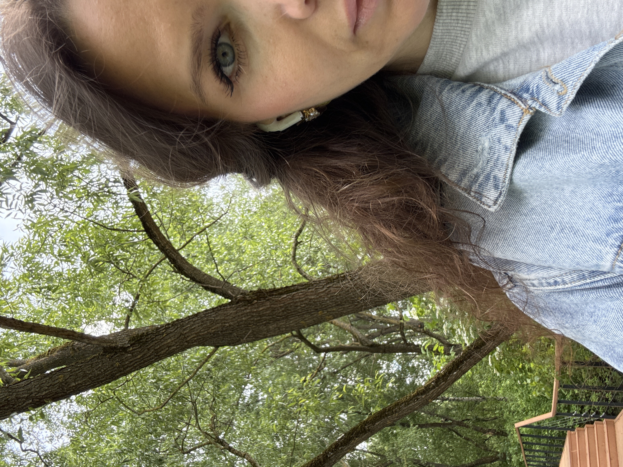 A selfie taken at an angle shows a person with fair skin, light-colored eyes, and brown hair with auburn highlights. The person is wearing a light gray top underneath a denim jacket, and an earring. The background consists of trees with green leaves and branches. A section of terracotta-colored stairs with black metal railings is visible. The location is Moscow, Russia.