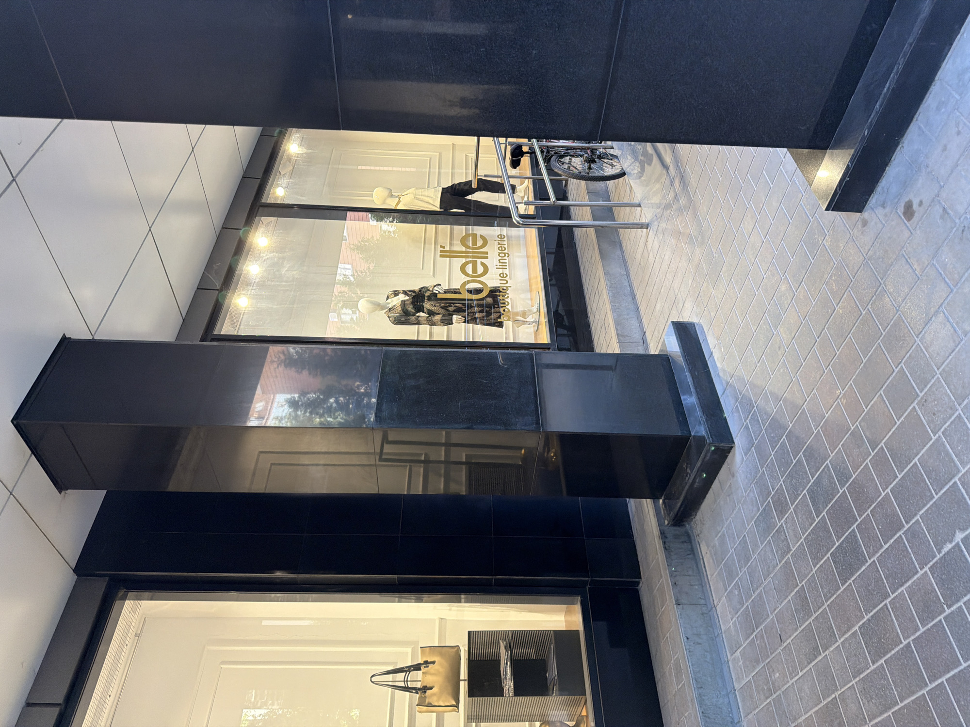 The image shows the exterior of a boutique called "belle lingerie" in Novaya Usman, Russia. The store features a large display window with mannequins dressed in clothing, and a beige handbag displayed on a black tiered stand. The shop's logo, "belle" and "boutique lingerie," is visible on the window. Outside the store, there is a bicycle parked near a metal railing, and the entrance area is paved with stone tiles. The building has a modern architectural style with dark stone pillars and a tiled ceiling.