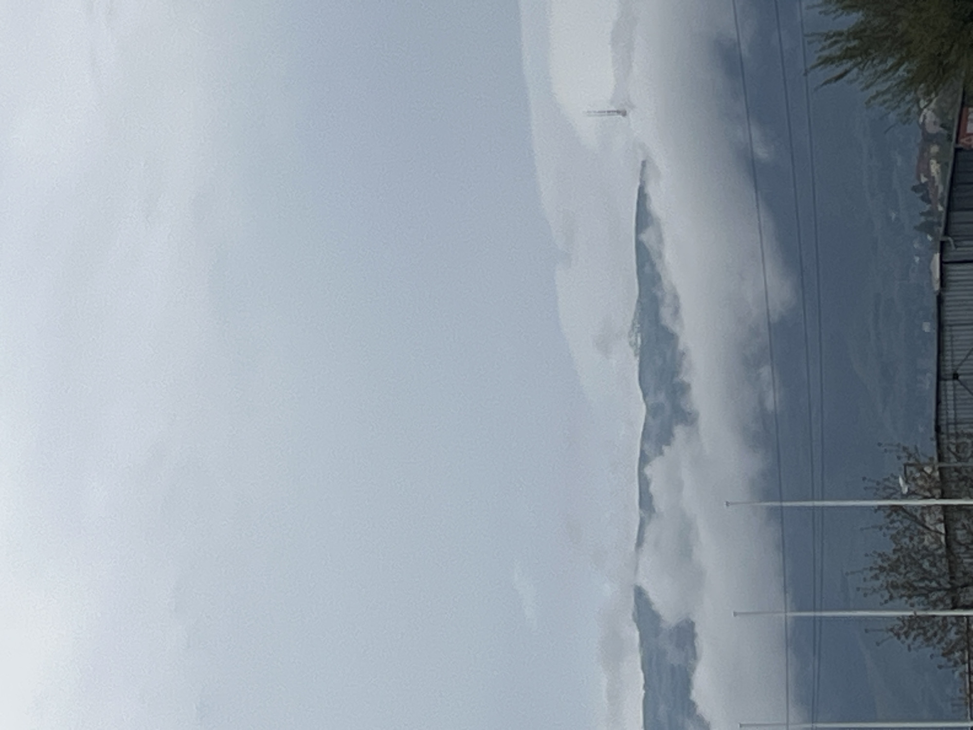 The image presents a view of what appears to be a mountain shrouded in clouds. Adjacent to the mountain are utility poles and some vegetation, possibly trees, indicating an inhabited area. The sky is overcast and grey. Given the general mountainous terrain, it is speculated to be in a city of Unknown, Unknown.