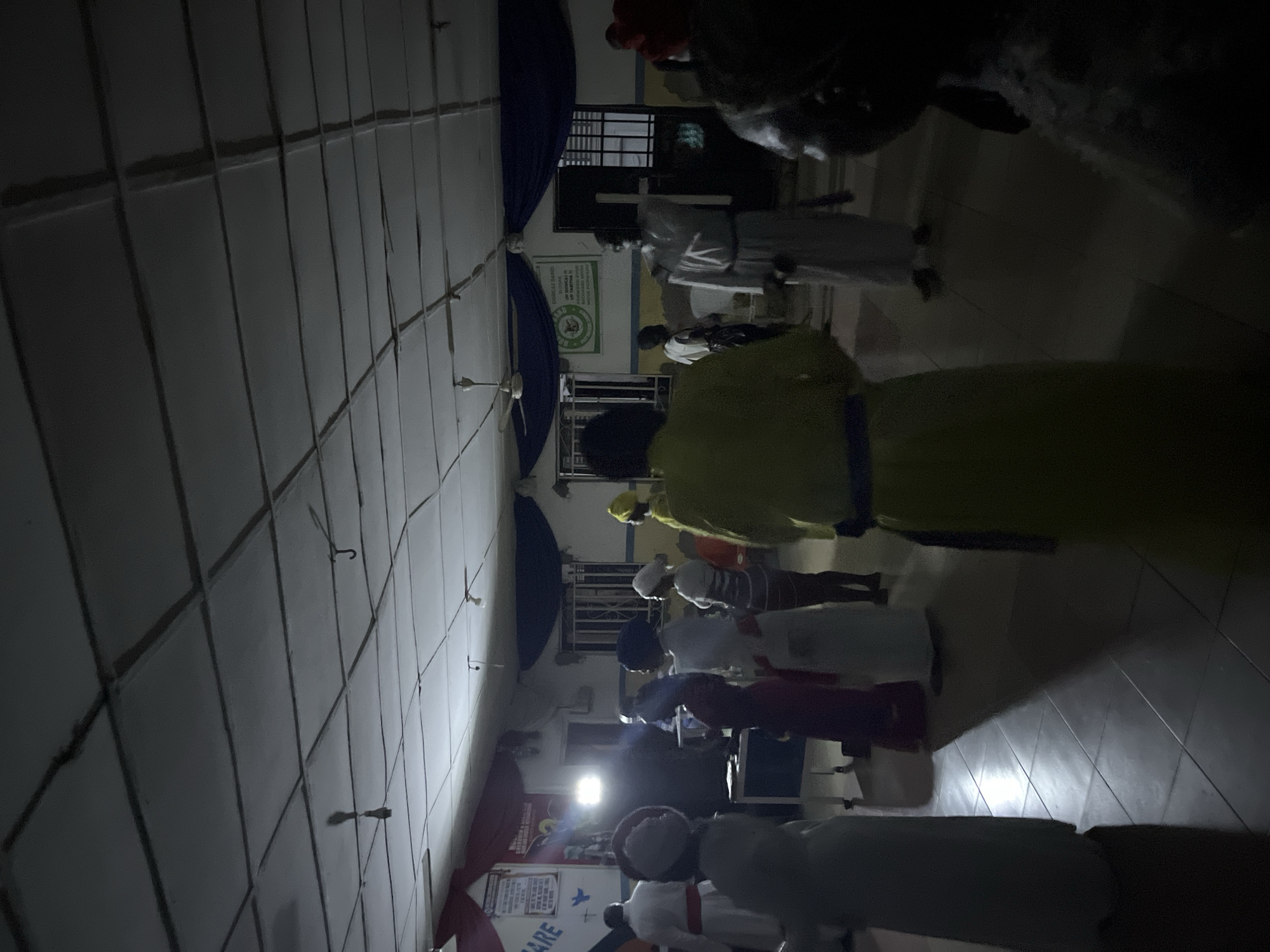 The image depicts an indoor gathering within a large hall, likely a community or religious center. The specific city and country are unknown. The room is dimly lit, primarily by artificial sources, with one particularly bright light emanating from the center-left, casting strong shadows on the floor. The ceiling is a drop-tile grid system with visible seams and some panels showing signs of wear or damage. Several ceiling fans and individual spotlights are mounted on the ceiling.
The walls are painted a light color and feature decorative elements, including red fabric drapes on the left side and dark blue drapes on the right side. Posters or banners are affixed to the walls; one on the left prominently displays the word "CARE," while another on the right, featuring a green circular emblem, includes text such as "DORCAS BAND," "SLOGAN," "UP DORCAS," and "MOVE FORWARD." Windows with protective metal grates are visible on the far walls. The floor is covered with light-colored square tiles.
Approximately 15 to 20 individuals are present, most of whom are wearing long robes or dresses. The attire varies, including white, yellow, red, and dark-colored garments, and many wear head coverings such as turbans or caps. Most individuals are standing, suggesting participation in an assembly or ceremony. Their faces are generally obscured due to the lighting conditions, angle, or position facing away from the camera.
Notable individuals include: a person on the far left in a white robe with a red sash; a person in the center-right wearing a white robe and holding a large, white cross; and in the immediate foreground, a person in a yellow robe with a blue sash standing with their back to the viewer, and another in a dark, patterned garment also facing away. A table with objects on it is visible in the center-left, near the bright light source.