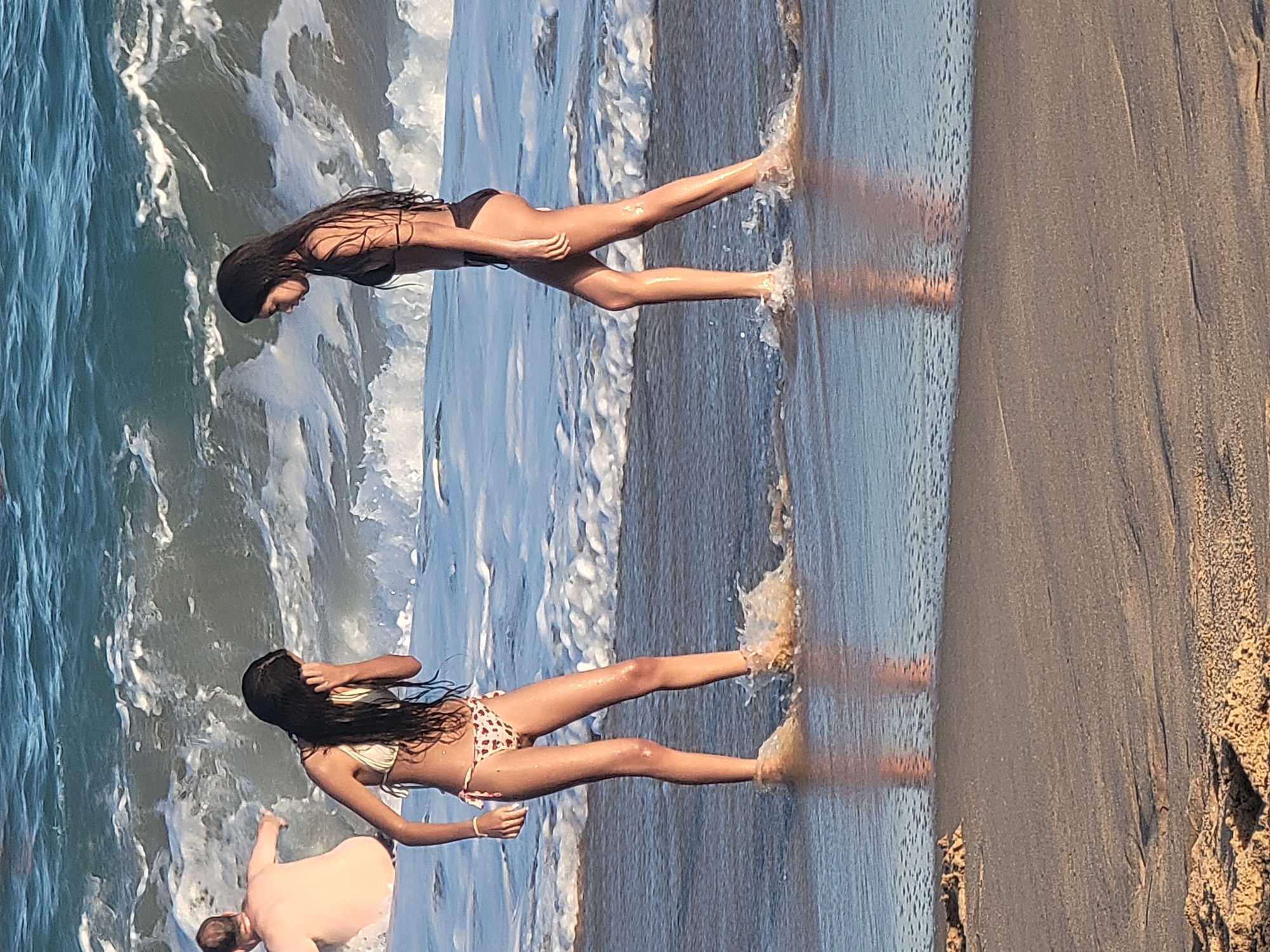 In Quarteira, Portugal, two young women in bikinis stand near the shoreline. The woman on the left, wearing a white and red patterned bikini, has her hand on her face. The woman on the right, in a black bikini, looks down. Waves are breaking around their legs, and a person in the background is partially submerged in the water. The scene takes place on a sandy beach under a bright sky.