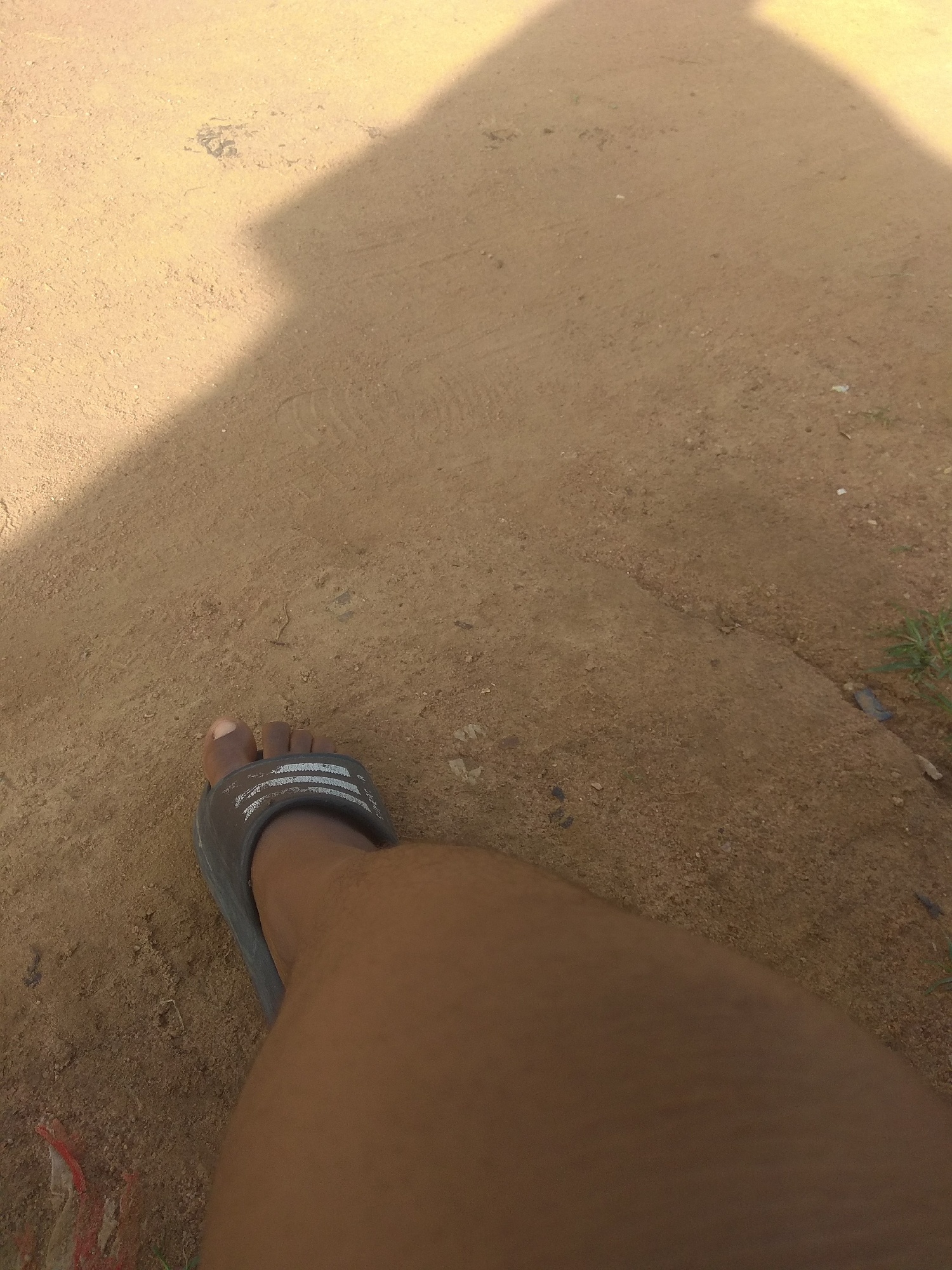 A low-angle, medium shot captures a person's left leg and foot, wearing a dark gray sandal with white stripes, positioned on sandy, uneven terrain. The sandal is positioned diagonally from the bottom left corner upwards and to the right. The foot is partially visible within the sandal. The ground is composed of light brown sand with scattered small pebbles and debris. A prominent shadow, cast by an unseen object, diagonally bisects the upper portion of the image from the top left to the middle right, creating a contrast between the sunlit and shadowed areas. The environment suggests an outdoor, possibly rural or undeveloped urban setting, consistent with the arid or semi-arid regions of Jalingo, Nigeria, where such sandy ground conditions are common. There is a hint of sparse green vegetation on the far right edge of the frame.