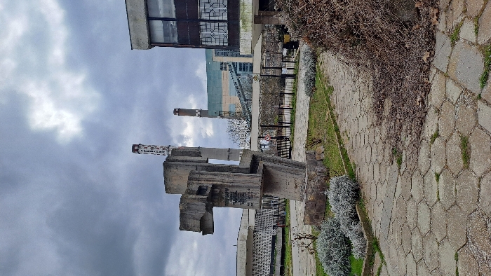 This image depicts an outdoor area in Ezerovo, Bulgaria, under an overcast sky. In the foreground, a pathway composed of hexagonal concrete pavers is visible, interspersed with patches of green grass. To the left of the path, a manicured garden bed contains several low-lying, silvery-green shrubs, while to the right, a large pile of dry, tangled brown vegetation rests on the path and adjacent ground.
Dominating the left midground is a large, weathered, abstract sculpture constructed from grey concrete blocks, mounted on a roughly textured stone base. Indistinct markings or graffiti are visible on its surfaces.
In the midground, beyond the pathway, a low, light-colored building with a patterned facade is partially visible to the left. To the right of the sculpture, a two-story building with a dark facade and grid-patterned windows stands. Between these structures, a covered outdoor area with multiple support columns and a flat roof is present, leading towards a fenced perimeter with what appears to be an access gate. Sparse, leafless trees are scattered behind the fence line.
The background is dominated by a large industrial complex, likely a power plant. The complex features a multi-level building with sections painted in shades of light green, blue, and beige. Two tall industrial chimneys are visible; the more prominent one on the left is cylindrical, white with red horizontal bands near its top, and has a pattern of rectangular openings along its shaft. A second, darker, and less distinct chimney stands to its right. A large, enclosed conveyor belt structure traverses the industrial site. The sky is largely grey and cloudy, with hints of brighter, diffused light in the upper right quadrant. No people or animals are present in the scene.