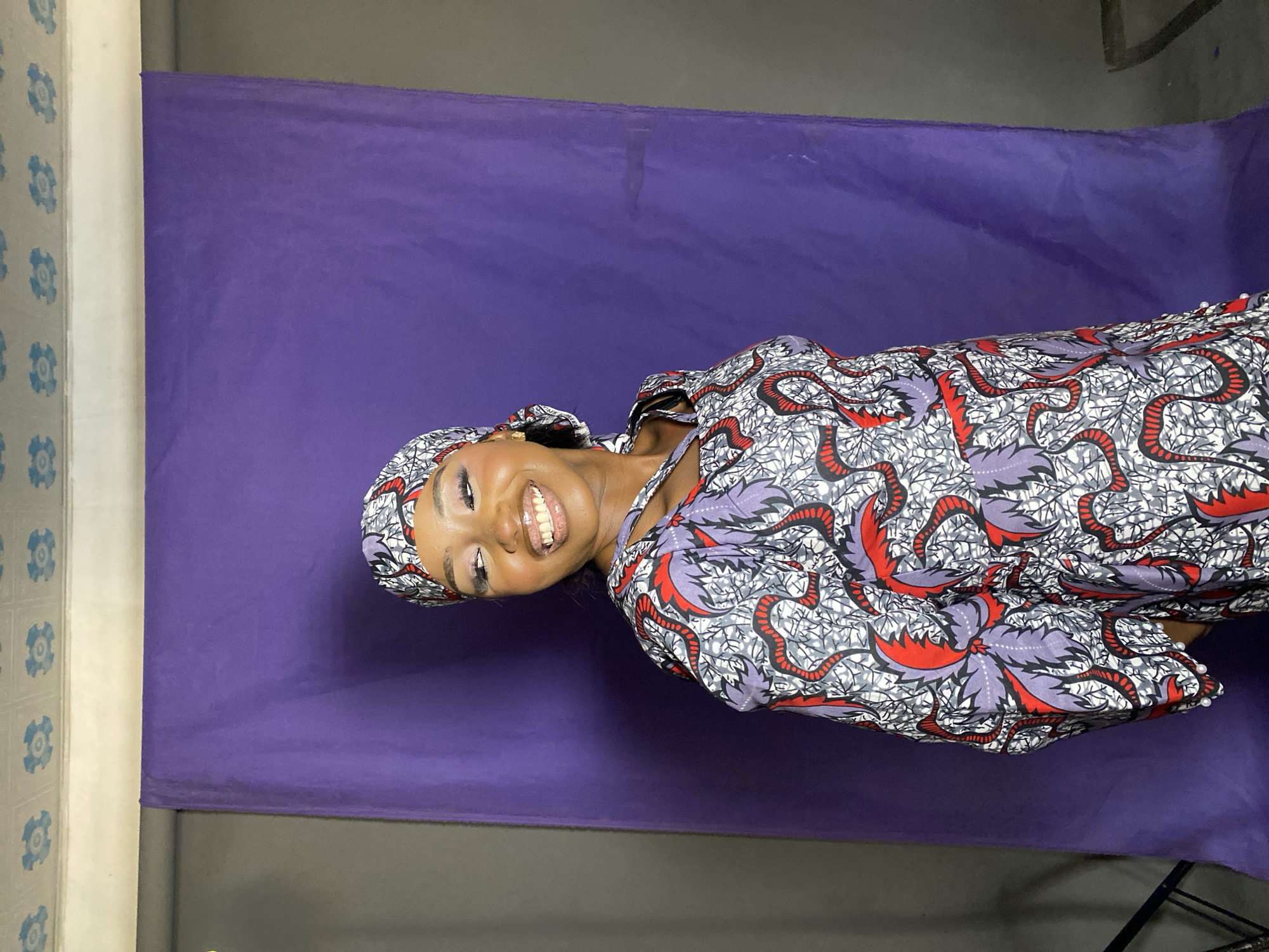 A woman is smiling at the camera while laying on a purple backdrop. She is wearing a head covering and a patterned robe-like garment in gray, white, red, and purple. The location is Gura Top, Nigeria. A patterned wall is visible to the left, and the backdrop sits atop a gray surface.