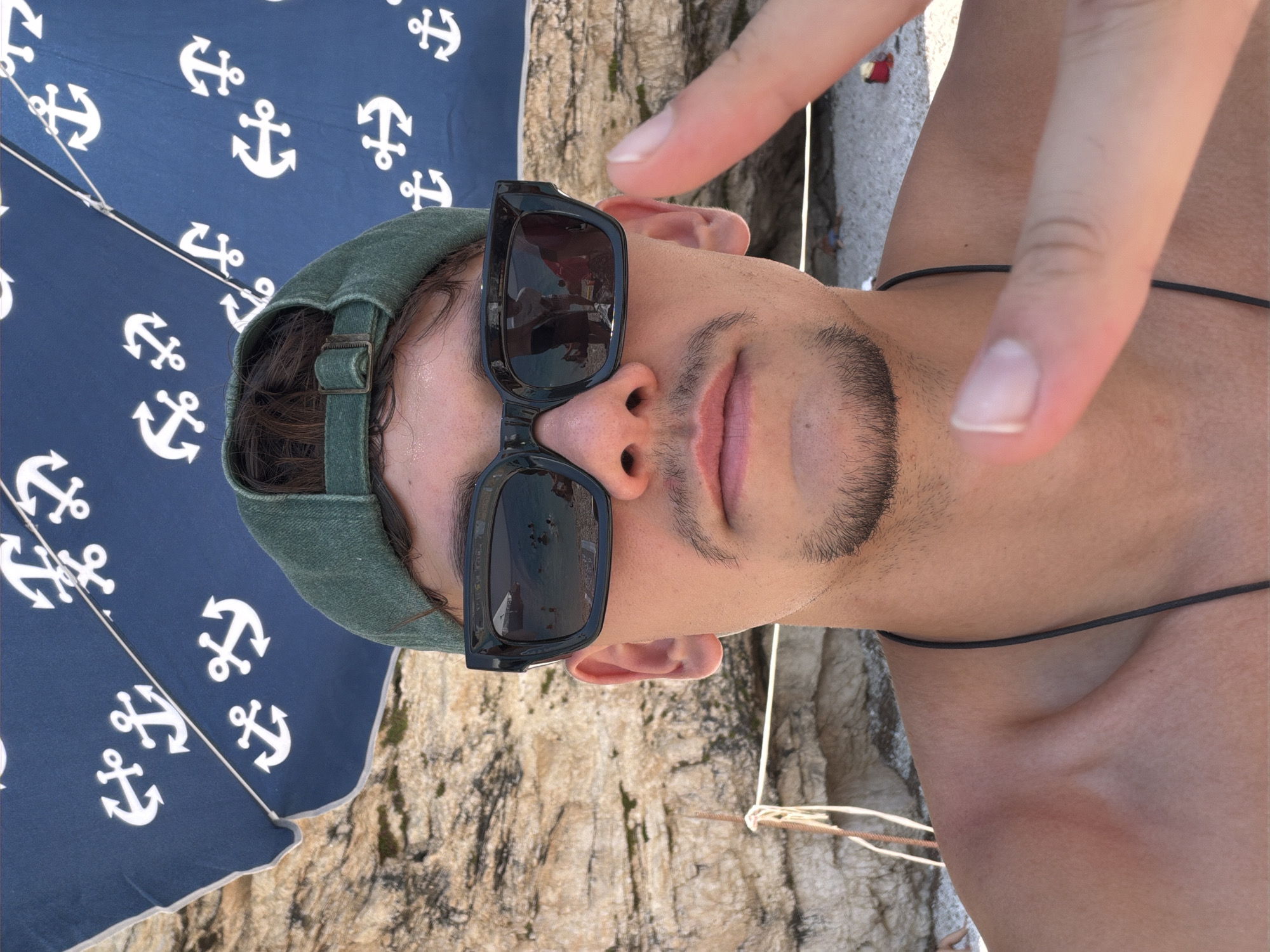 A young man with dark hair, a goatee, and a mustache poses for a selfie on a beach in the Municipal Unit of Apollonioi, Greece. He is wearing a green baseball cap and black sunglasses. The sunglasses reflect a beach scene with people swimming in the water. Behind him, a blue umbrella with white anchor patterns is visible. A rocky cliff face and a white rope are also in the background. He is holding up his right hand in a peace sign. The image is taken in natural daylight.