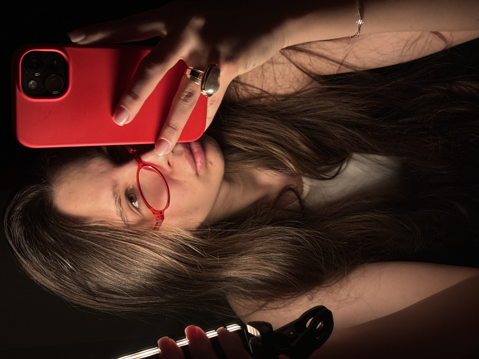 A young female, appearing to be in her twenties, is captured in a close-up portrait, likely a selfie. She possesses long, wavy brown hair that falls over her shoulders and part of her chest. She wears small, oval-shaped red-framed glasses. Her gaze is directed towards the viewer, with a neutral expression. She is wearing a light-colored top, possibly white or cream.
Her right hand holds a red smartphone encased in a red protective cover, positioned to obscure the lower right portion of her face. The phone exhibits a triple camera system on its rear. On the ring finger of her right hand, she wears a prominent ring featuring a gold-colored band and a large, dark, oval-shaped setting. A delicate bracelet with a small, reflective element is visible on her left wrist.
Her left hand, positioned on the left side of the frame, holds a strip of vertical LED lights, attached to a black clamp. These lights emit a warm, bright glow, serving as a primary light source for the scene. The background is uniformly dark, providing no discernible environmental details. The lighting creates strong highlights on her face, hair, and the illuminated objects, with significant shadows elsewhere.
The image provides no visual information to determine a specific location within the city of Krasnodar, Russia.