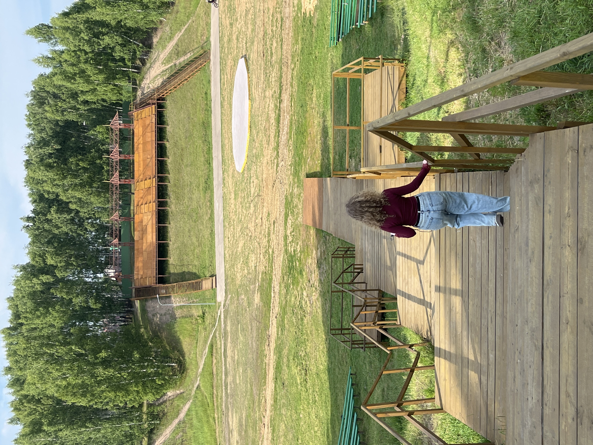 A person with curly hair, wearing a burgundy top and light blue jeans, walks down a wooden staircase in an outdoor amphitheater complex. The staircase, constructed of weathered wood, has a railing on the right side. In the background, a tiered wooden amphitheater with rusty metal seating is visible, backed by a dense forest of birch trees. A circular white platform, possibly a stage, is situated in the grassy area in front of the amphitheater. Green bleacher seats are arranged to the left of the amphitheater. The ground is covered in grass and dirt paths. The scene appears to be in Zelenodol'sk, Russia, likely an outdoor recreational or cultural venue.
