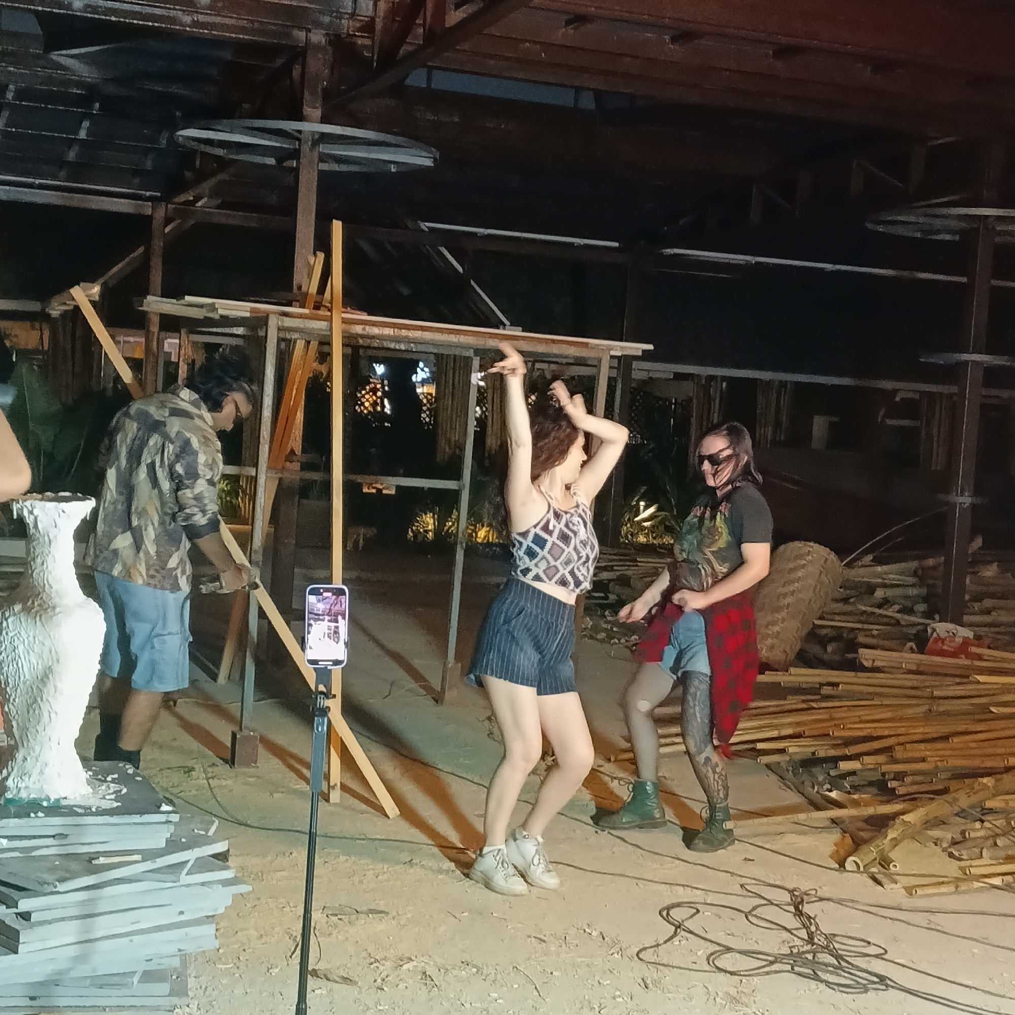 The image captures a scene in an unfinished or under-construction space, possibly in Panaji, India. The setting appears to be a large open-air structure with a dark, wooden framework above, suggesting it may be a building site or a covered outdoor area. There are three individuals visible: a person on the left, wearing a camo-patterned shirt and blue shorts, appears to be working with wood, near a phone on a stand, recording him. Two women in the foreground are engaged in a dance, with one positioned closer to the camera and wearing a patterned crop top and striped shorts. The other, slightly behind, is wearing a dark t-shirt and a red plaid shirt tied around her waist. The environment is cluttered with building materials, including piles of wood. There are also stacked tiles or slabs visible near the worker. Lighting in the scene seems dim, with the primary source coming from the phone’s light.