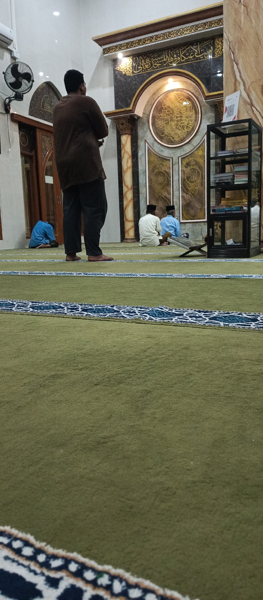 The image depicts the interior of a mosque in Arjasa, Indonesia. The foreground is dominated by a green carpet with a decorative blue and white border. In the middle ground, a person stands with their back to the camera, wearing a brown shirt and dark pants. To the left, a person in a blue shirt is seated on the floor. Further back, two individuals in white and blue shirts respectively, both wearing skullcaps, are seated on the floor facing the Qibla wall. The Qibla wall features ornate Arabic calligraphy within circular and rectangular frames. A black glass shelving unit holding books is visible to the right of the Qibla wall. A fan is mounted on the wall to the left of the scene. The lighting suggests it is indoors.