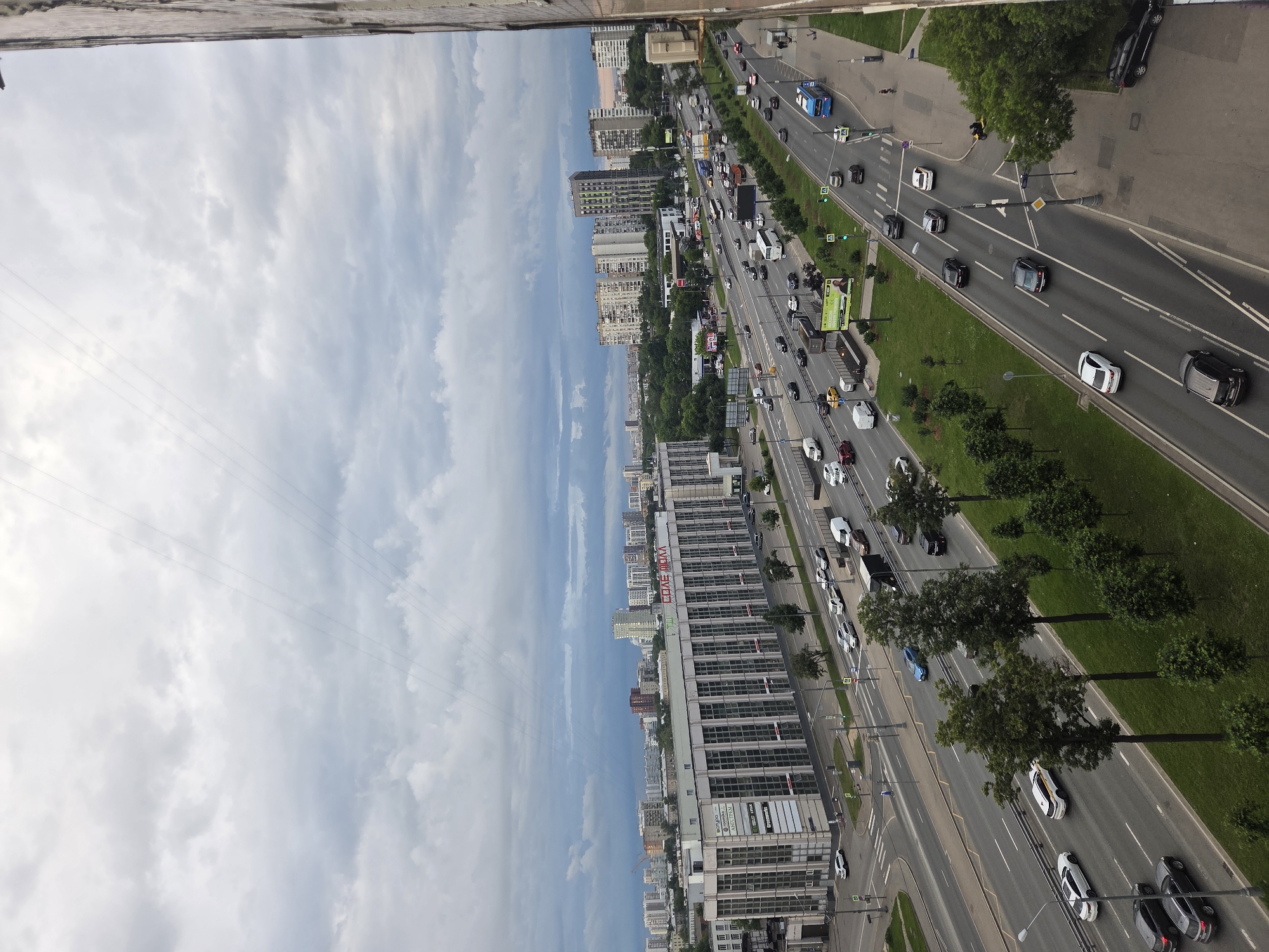 This is a high-angle, slightly tilted, medium shot of a busy, multi-lane highway in Moscow, Russia, on a cloudy day. The highway is flanked by numerous tall residential and commercial buildings, and lush green trees line the roadside. Cars, buses, and trucks are actively navigating the roadway in both directions, indicating a period of moderate to heavy traffic.
The buildings are a mix of modern high-rises and older, more uniform apartment blocks. Some of the buildings have visible signage. On one of the larger buildings, the red text "КОНЕ ЧОМ" is clearly legible. Another building displays text such as "ПРОМЫШЛЕННЫЙ", "СТРОИТЕЛЬСТВО", and "АВТОКОМПЛЕКС". The sky is predominantly overcast with scattered clouds, suggesting it is daytime, likely afternoon. The overall scene depicts an urban environment in motion, with the infrastructure of a large city and the constant flow of vehicular traffic. There are no people visibly identifiable in the scene, only the vehicles and the urban landscape.