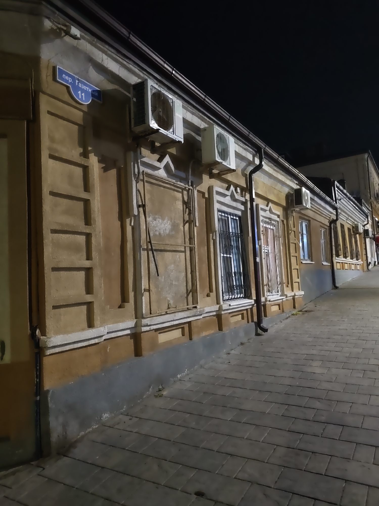 The image shows a nighttime view of a street in Rostov-on-Don, Russia. The building on the left is a light brown, two-story structure with decorative architectural elements. A street sign for "пер. Газетный 11" (Gazetny Lane, 11) is visible on the upper part of the building. Multiple air conditioning units are mounted on the building's facade. The windows on the first floor are covered with metal grates or shutters. The sidewalk in front of the building is paved with gray interlocking bricks. The scene appears to be quiet and undisturbed.