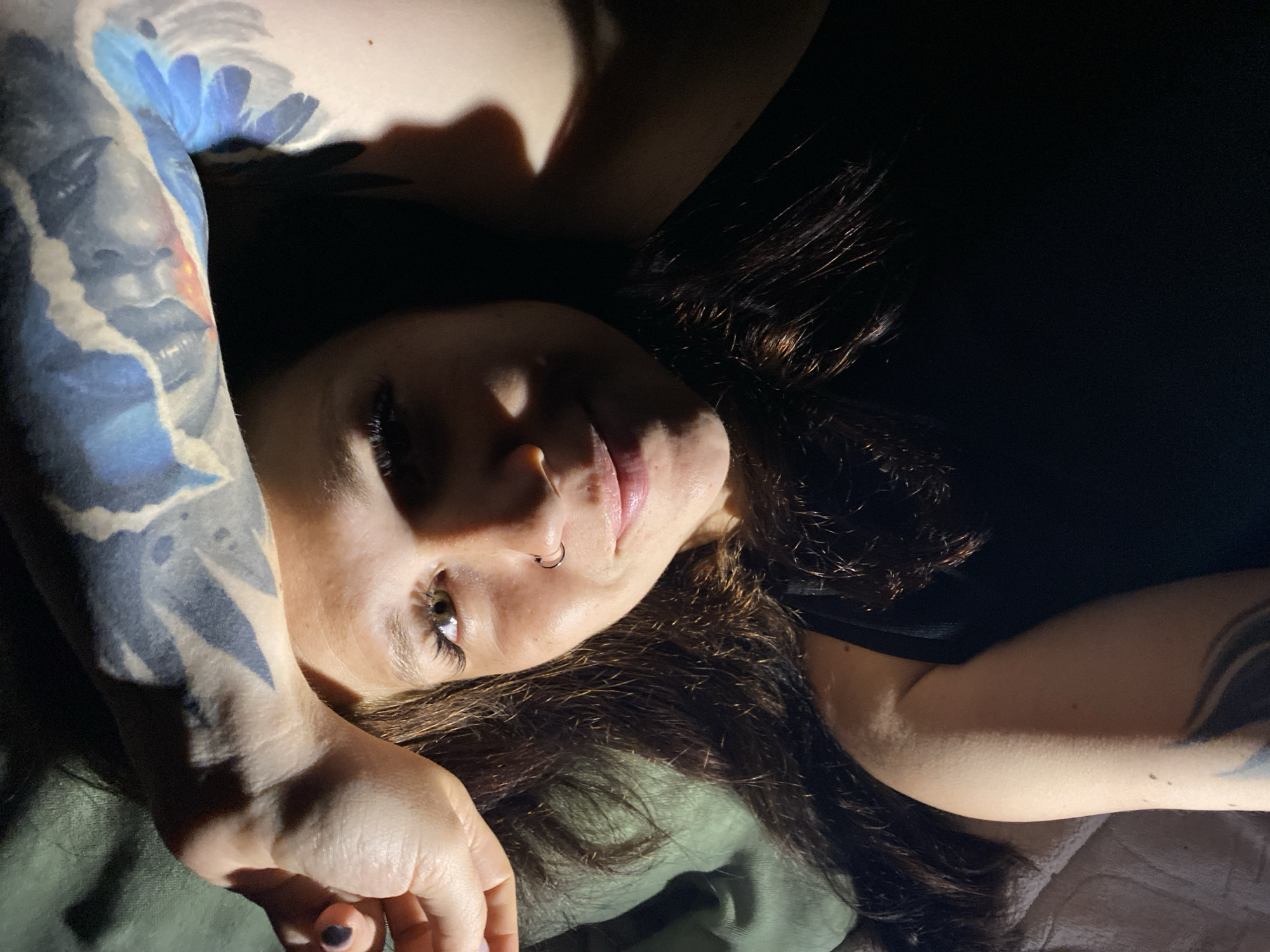 A close-up, overhead shot shows a woman lying down in low lighting, with her arm raised to cover part of her face. She has long, dark, wavy hair, and is wearing a black tank top. She has a nose ring and large eyelashes, and her skin has freckles. There is a tattoo on her arm, partially obscured by her forearm. The background is dark, with a hint of green fabric on the left. The image was taken in Chaikovsky, Russia.