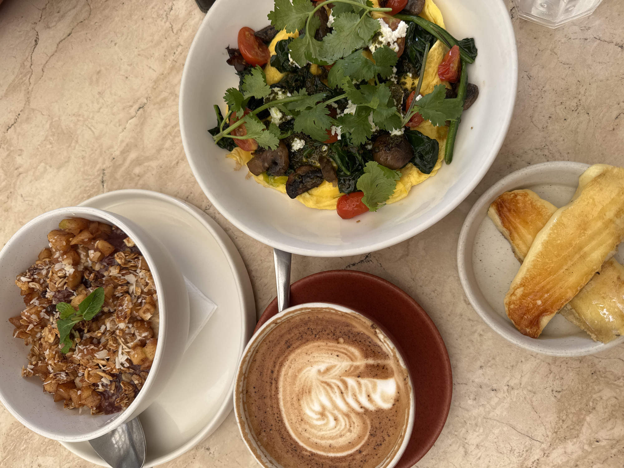 An overhead shot captures a vibrant breakfast or brunch spread laid out on a light-colored, veined marble or stone tabletop. The scene is brightly lit, suggesting daytime.
In the upper center, a white bowl holds a savory dish, likely an omelet or scrambled eggs. It's generously topped with sautéed spinach, dark mushrooms, bright red halved cherry tomatoes, crumbled white cheese (possibly feta), and a vibrant green sauce or pesto, all garnished with abundant fresh cilantro leaves.
To the bottom left, another white bowl contains a sweet breakfast item. It features a mixture of what appears to be stewed fruit (like apples), granola, and shredded coconut, with a small fresh mint sprig as a garnish. A spoon rests in the bowl, which sits on a matching white saucer.
A cup of coffee, likely a latte or cappuccino, is positioned at the bottom center, showcasing detailed latte art on its foam. The white cup has a brown rim and rests on a reddish-brown saucer.
On the right side of the frame, a small, speckled light gray ceramic plate holds three pieces of golden-brown, possibly fried or baked dough, which could be plantains, pastries, or bread sticks. A portion of a clear, textured glass is visible in the top right corner.
The overall arrangement suggests a curated and enjoyable dining experience, likely at a cafe or restaurant specializing in brunch. No text is visible in the image.