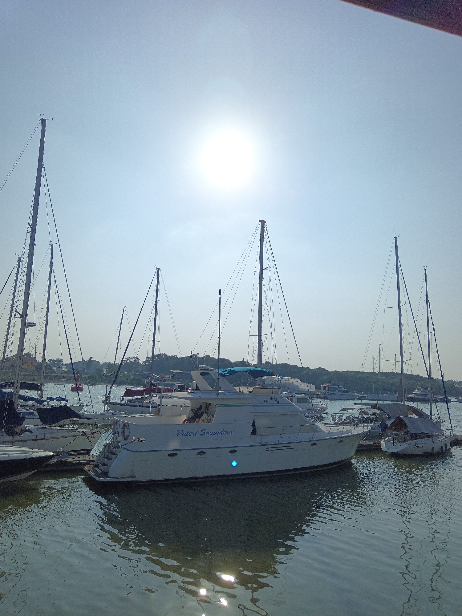The image shows a sunny day at a harbor in Klang City, Malaysia. Several sailboats and yachts are docked in the water. The sun is shining brightly in the sky, casting a glare on the water. The boats are of various sizes and designs, with some having tall masts and sails. The foreground is dominated by the water, with ripples reflecting the sunlight. In the background, there is a treeline and some buildings, suggesting a coastal or riverside location. The overall scene depicts a peaceful maritime setting.