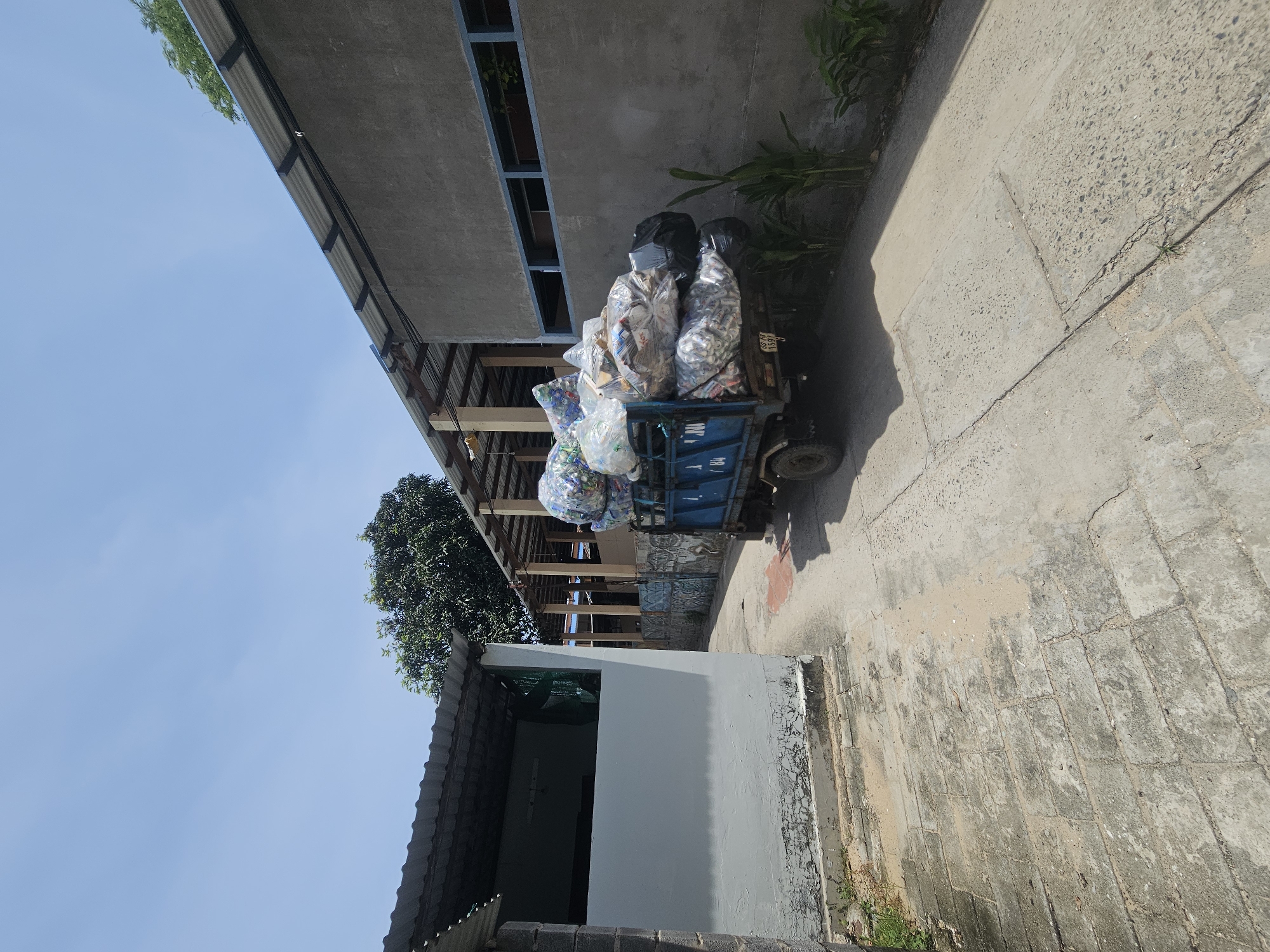 A light blue truck is parked on a concrete path in a yard with several industrial buildings and a large tree under a clear blue sky. The truck is loaded with large, clear plastic bags filled with recyclable materials, primarily plastic bottles. The truck is positioned in the middle ground of the image, facing towards the right. To the left of the truck, a white building with a corrugated metal roof is visible. To the right, a grey building with a corrugated metal roof and a blue-trimmed window is present. The ground is a mix of concrete and brick paving, with some dirt and sand scattered about. Plants are visible growing along the base of the grey building. The scene appears to be a recycling or waste management facility. The location is identified as Phu Thuy Ward, Vietnam.