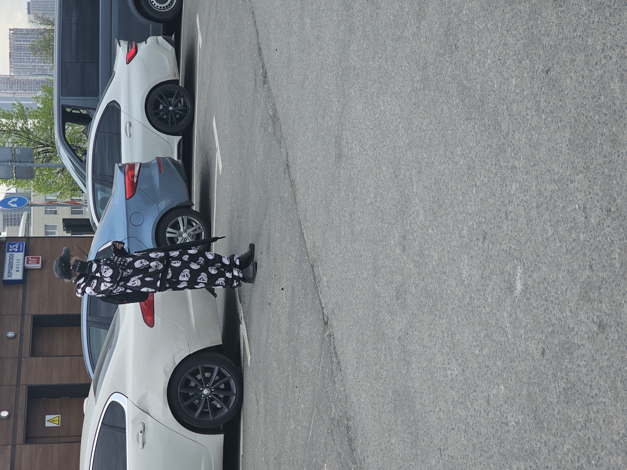 A person dressed in skull-patterned clothing stands next to a white car parked on a street in Moscow, Russia. The person is wearing a hat, a mask covering part of their face, and black shoes. The sign reads "Khoshchevskoe Shosse 35". In the background are a blue car, a white car, and a gray van. Buildings and trees are visible in the distance. The area appears to be a parking spot alongside a building.