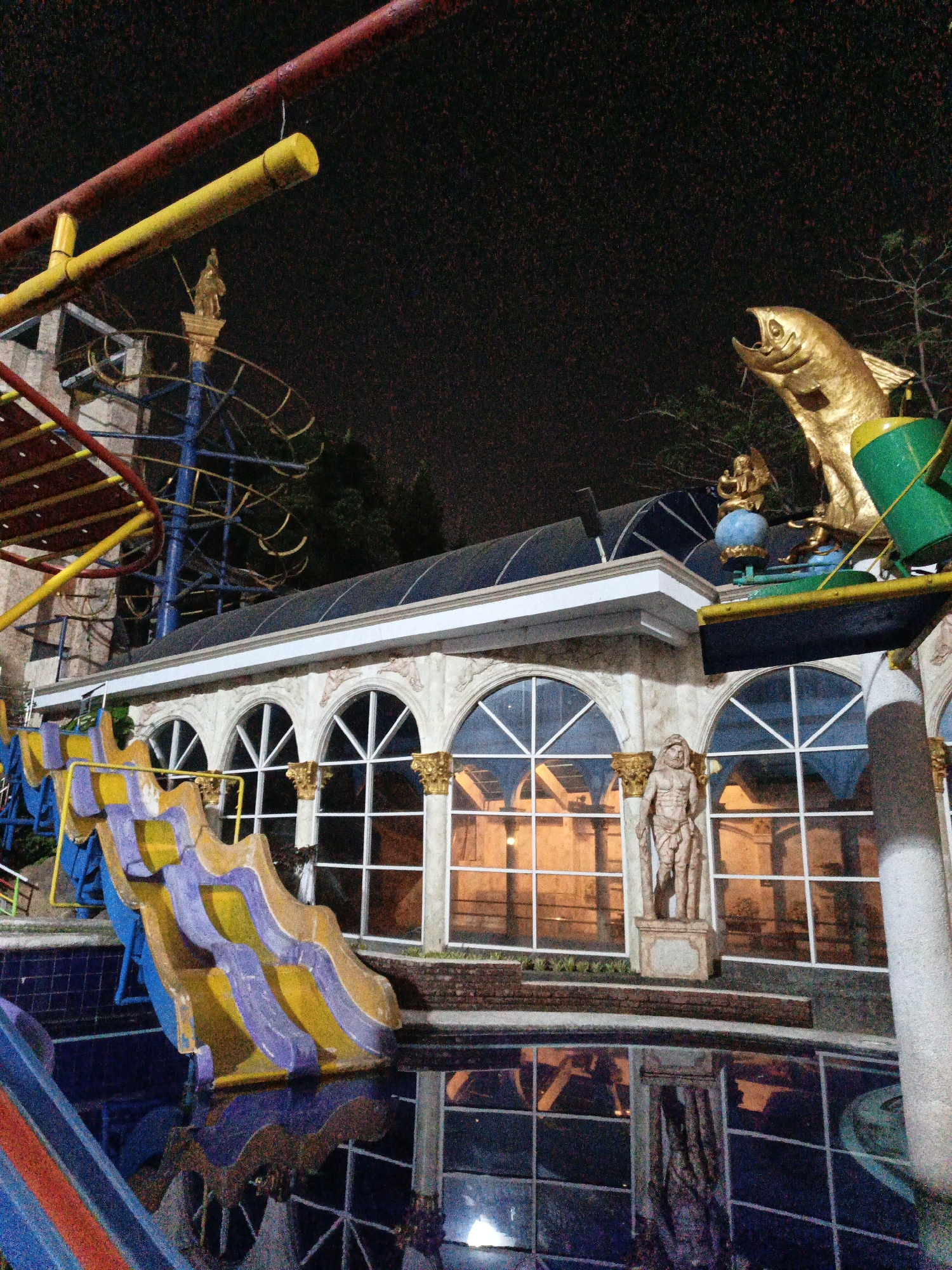 The image depicts an outdoor water park or amusement park area at night in Cibogo, Indonesia. Dominating the scene is a brightly colored water slide with purple, yellow, and blue sections, ending in a swimming pool whose surface reflects the surrounding lights and structures. Above and behind the slide is a building with arched windows and decorative gold columns. A statue of a muscular, robed male figure stands on a pedestal in front of the building. To the right of the building, a large, golden fish sculpture with its mouth open is prominently displayed, along with a green and black object that appears to be part of an attraction. In the background, a dark sky is visible, with trees and parts of a ride structure, possibly a Ferris wheel, partially obscured. Red and yellow pipes, likely part of another ride or structure, are also visible in the upper left of the frame. The overall impression is of a festive, illuminated recreational space.