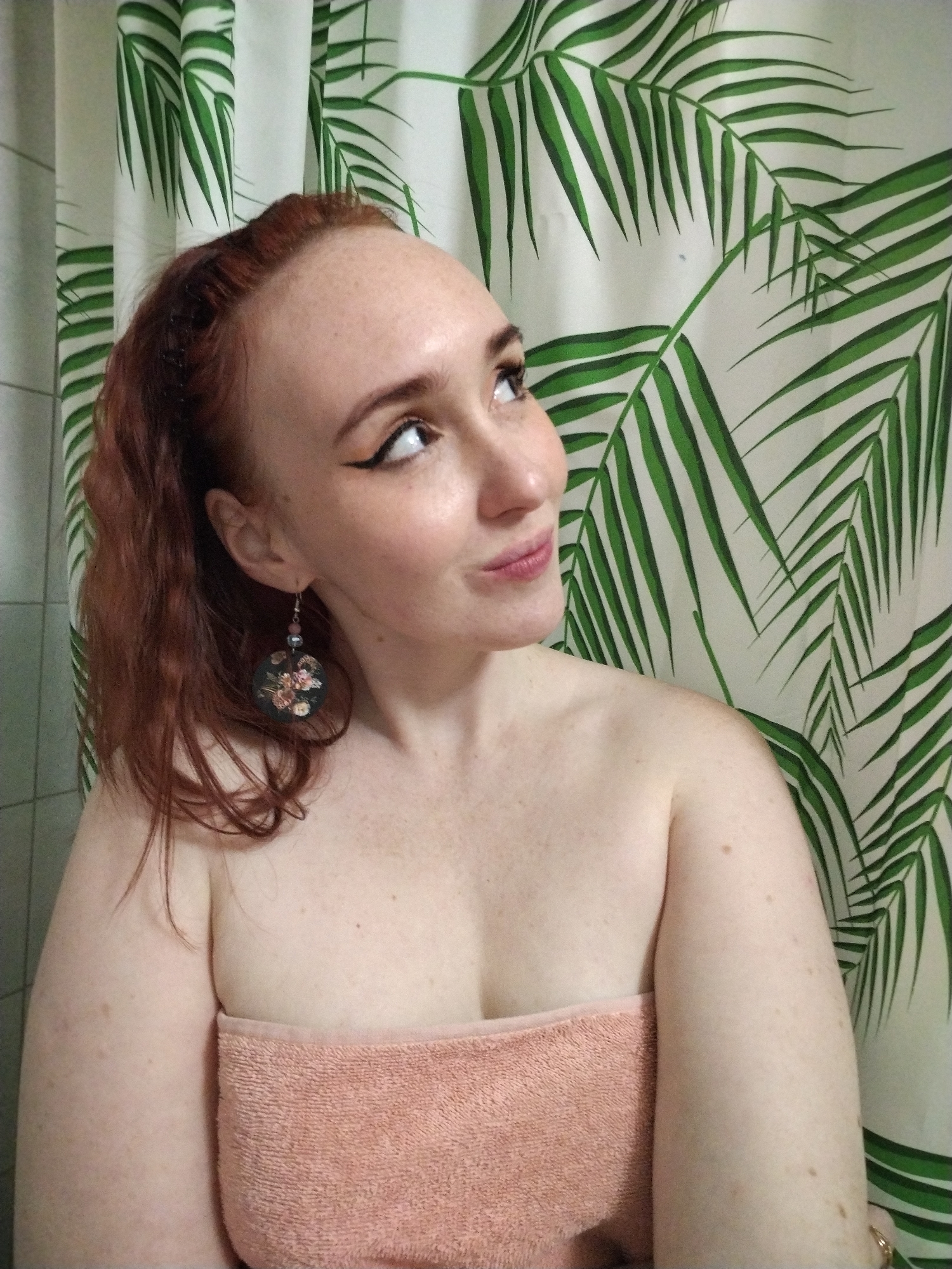 A red-haired woman with fair skin and freckles is shown from the chest up, wrapped in a peach-colored towel. She is wearing eyeliner and an earring with a floral design. Her gaze is directed upward and to the right. Behind her is a shower curtain with a pattern of green tropical leaves on a white background. The setting appears to be a bathroom. There is no information to associate the scene with the city of Izhevsk, Russia.