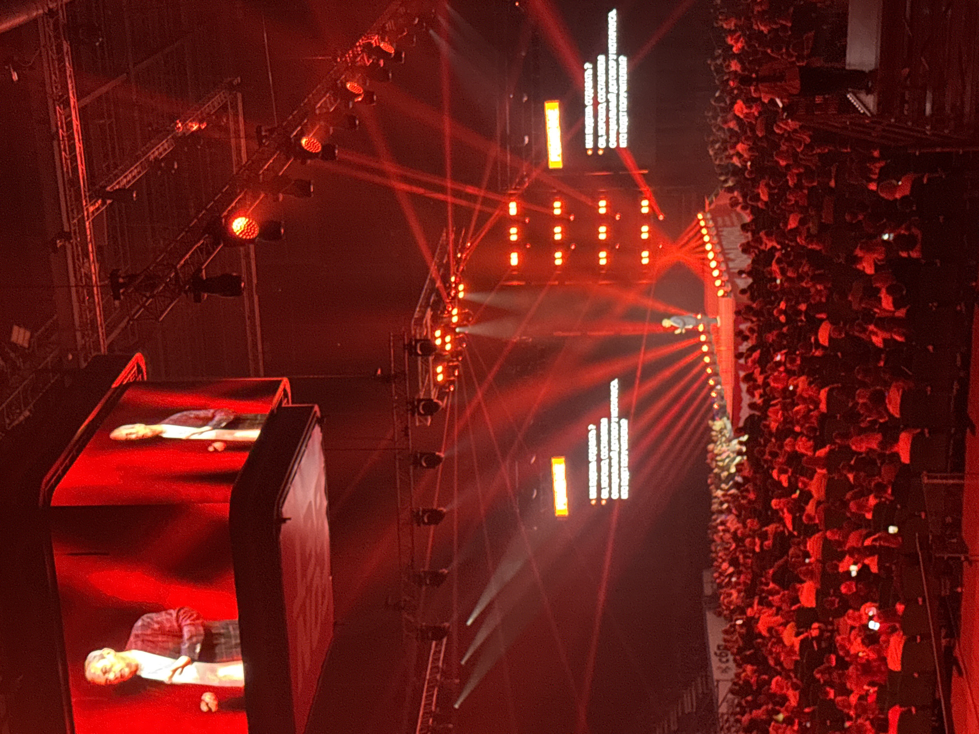 The image depicts a large-scale event, likely a conference or performance, taking place in the city of Ufa, Russia. The scene is dominated by red lighting, creating a dramatic atmosphere. On a large stage, two individuals are projected onto a screen, appearing as if they are standing there. One is wearing a patterned jacket over a white shirt, and the other, further to the right, is in a similar outfit. Spotlights and stage lighting are prominent, casting beams of light across the venue. Below the stage, a vast audience is seated in rows, their faces illuminated by the red glow. Some audience members are holding up their phones, likely recording or taking pictures. The overall environment suggests a modern indoor venue, possibly a stadium or exhibition center, equipped for large gatherings. The text visible on the screen is in Russian and appears to be related to legal matters or contracts, indicated by phrases like "КАКОЙ ДОГОВОР", "СОСТАВИТЬ", and "НОТАРИУС".