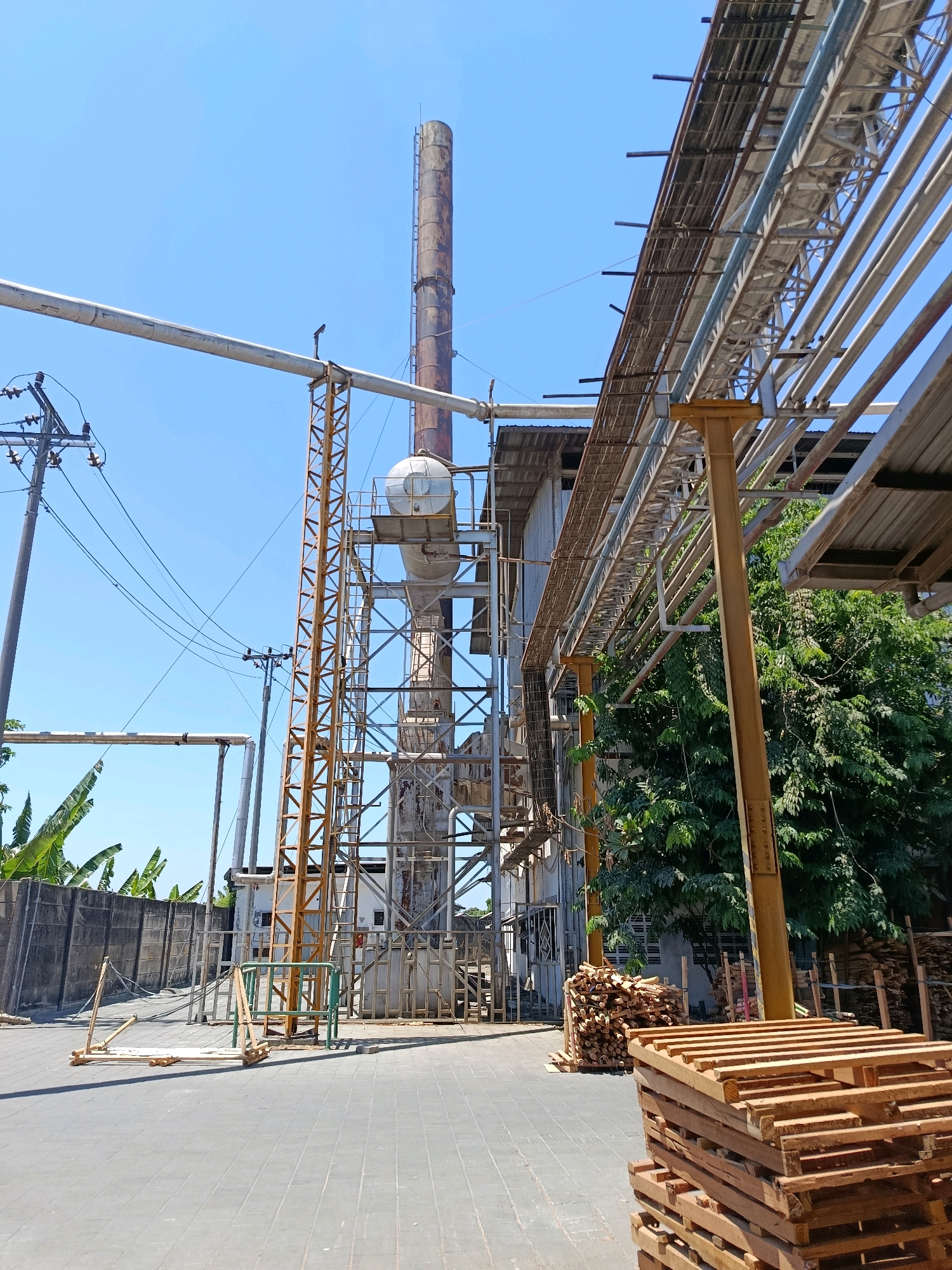 In Kembangarum, Indonesia, an industrial area is depicted under a clear blue sky. A tall, weathered smokestack dominates the center of the frame, connected to a complex network of pipes and metal structures. To the right, a yellow support beam holds up an overhead conduit filled with pipes, stretching towards a cluster of green trees. In the foreground, stacks of wooden pallets and lumber are neatly arranged on a paved ground surface. A concrete wall with banana trees visible behind it lines the left side of the scene, and a utilitarian building with a corrugated roof is situated behind the smokestack. Electrical poles and wires are also present, adding to the industrial ambiance.