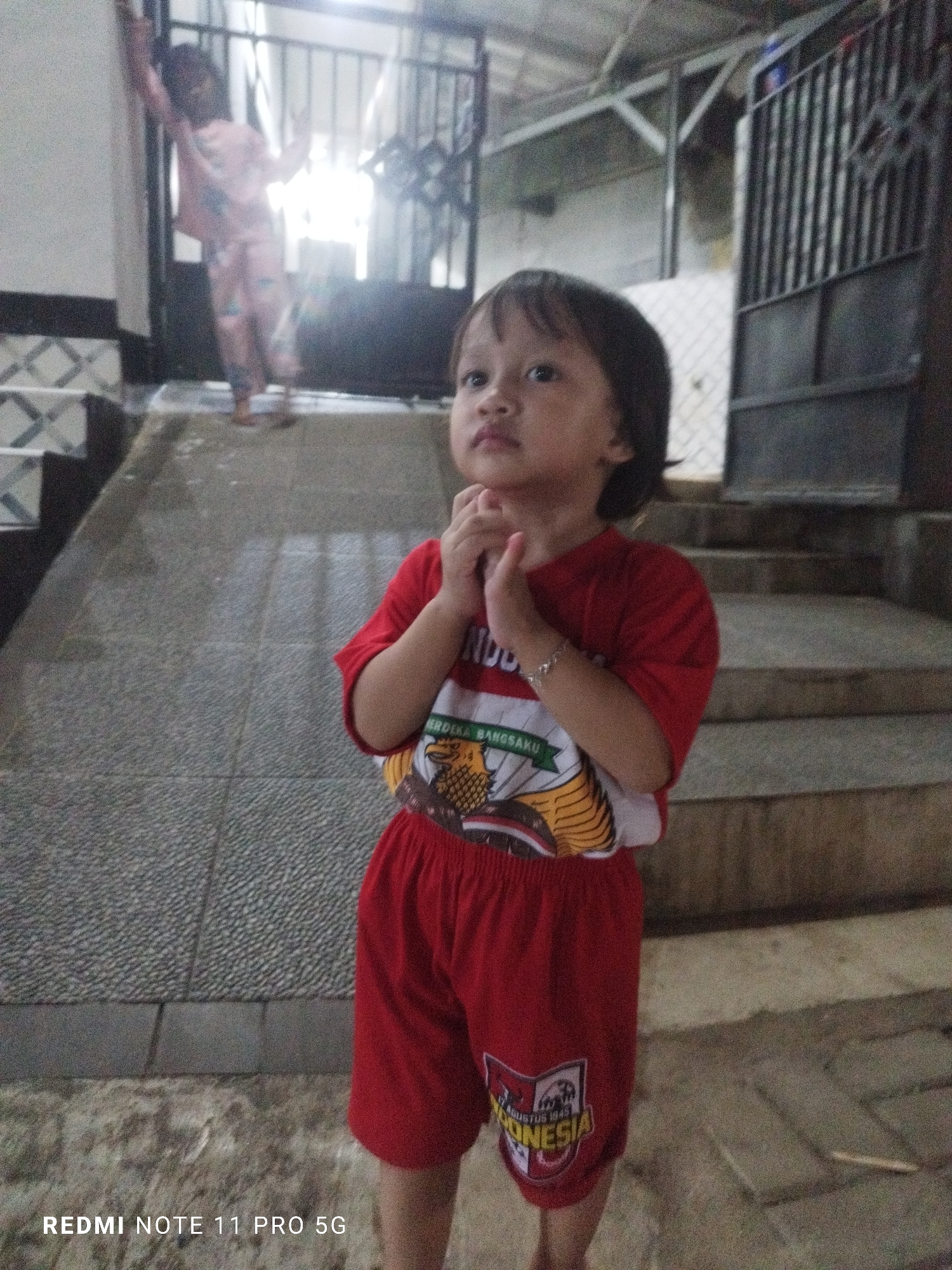 A young child, appearing to be of Southeast Asian descent and possibly Indonesian, stands on a paved walkway in Kaduhejo, Indonesia. The child is wearing a red t-shirt with a graphic print and red shorts. Their hands are clasped together in front of their chest. In the background, another child is visible, wearing pink pajamas and standing near a metal gate. The environment suggests an outdoor setting with stairs and a building visible. The image has a watermark at the bottom left corner reading "REDMI NOTE 11 PRO 5G".