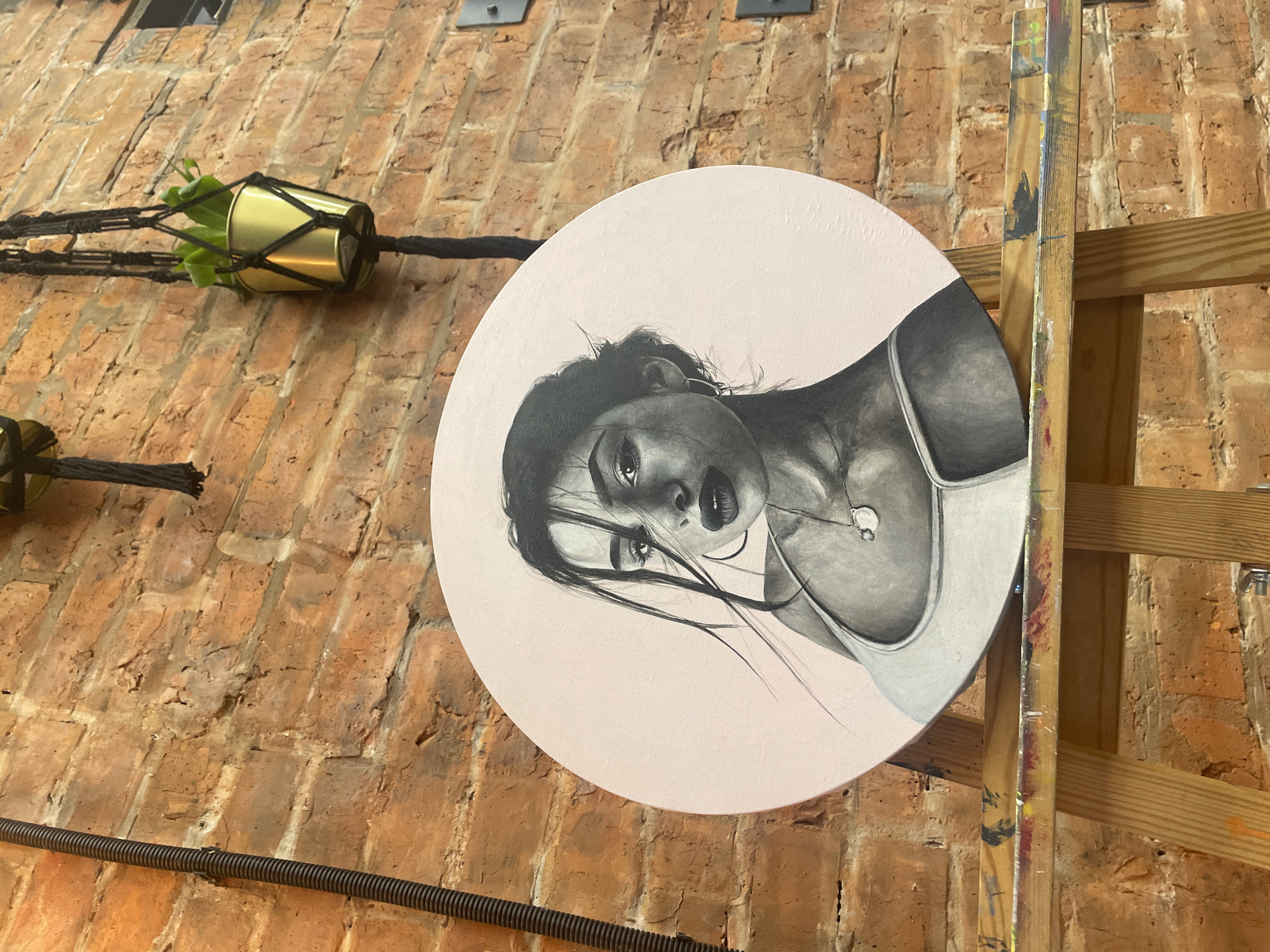 This is a photograph taken at an indoor location, likely an art studio or gallery, with a brick wall as the primary background. A large, circular painting is displayed at an angle, resting on a wooden easel. The painting is a black and white portrait of a woman, rendered in a realistic style with dramatic shading. Her gaze is directed downwards and to the side, with her hair falling across her face. She is wearing a tank top and a necklace with a pendant. The background of the painting is a light pink hue.
In the upper left portion of the image, a small potted plant in a gold-colored planter hangs from a macrame hanger attached to the brick wall. Another similar planter is partially visible further down. The brick wall shows signs of age and texture, with visible mortar lines. The wooden easel has paint splatters on it, suggesting it's actively used by an artist.
The lighting appears to be artificial, casting shadows that highlight the texture of the brick and the details of the painting. The overall mood is artistic and perhaps a little contemplative, evoked by the portrait's expression and the studio setting. There is no visible text in the image.