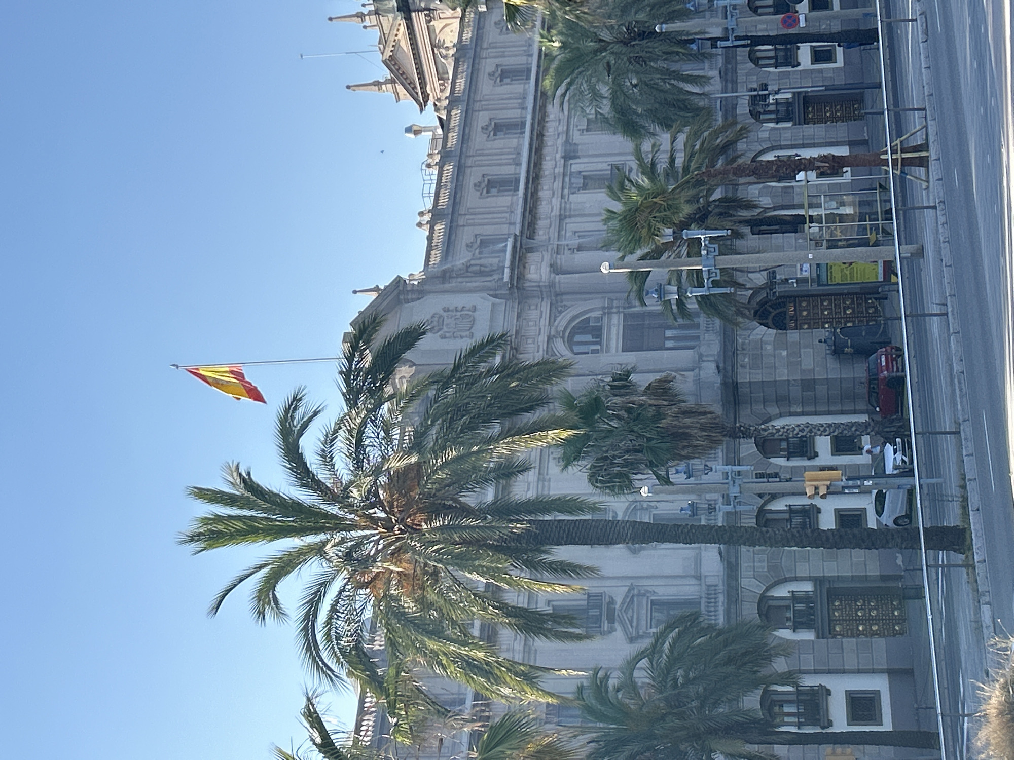 The image captures a sunny day in Barcelona, Spain, with the iconic building of the Port Authority of Barcelona serving as the central subject. A Spanish flag flies proudly from a flagpole atop the building. Several palm trees are in the foreground, their fronds reaching towards the clear blue sky. The Port Authority building exhibits neoclassical architecture with grand columns, ornate detailing, and arched windows. In front of the building, a street is visible with traffic signals, streetlights, and a few parked cars, including a white SEAT and a red car. A bus stop shelter is also present. The scene is set against a backdrop of the bright blue sky, suggesting a warm and pleasant day.