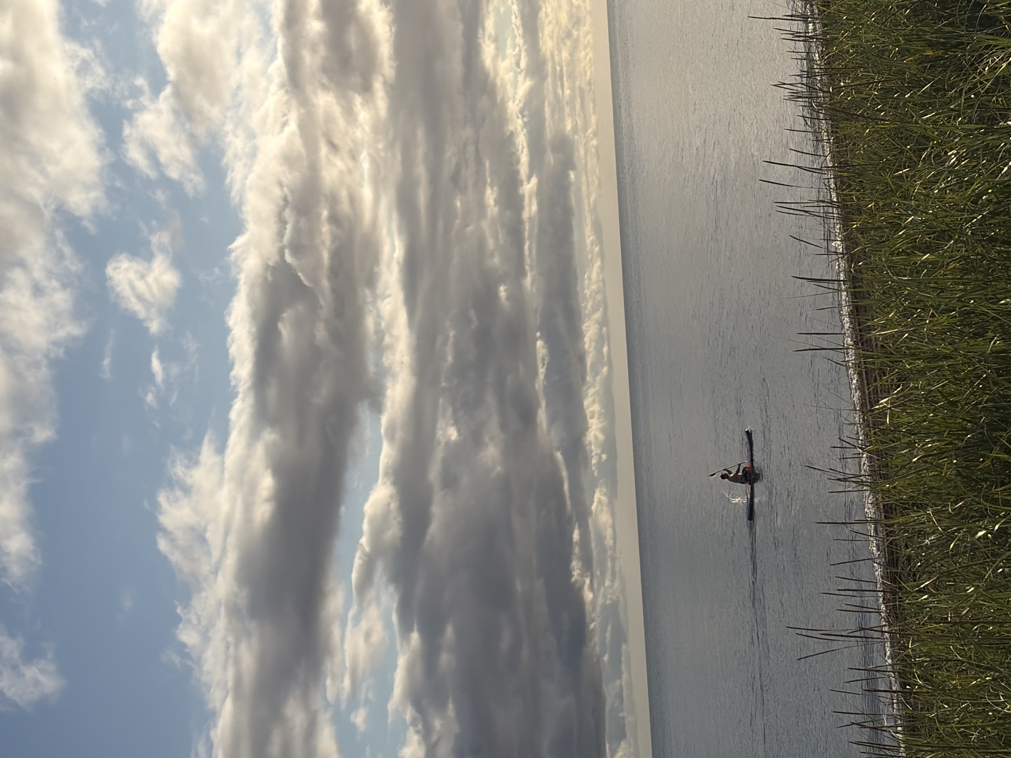 The image depicts a serene natural scene with a body of water, likely a lake or sea, in the foreground. Tall, green grass lines the shore, with the water extending to the horizon. The sky above is filled with dramatic, layered clouds, with patches of blue sky visible through them. In the middle ground, a solitary person is seen on a paddleboard, actively paddling and creating a small splash. The overall impression is one of tranquility and outdoor recreation in a natural setting.