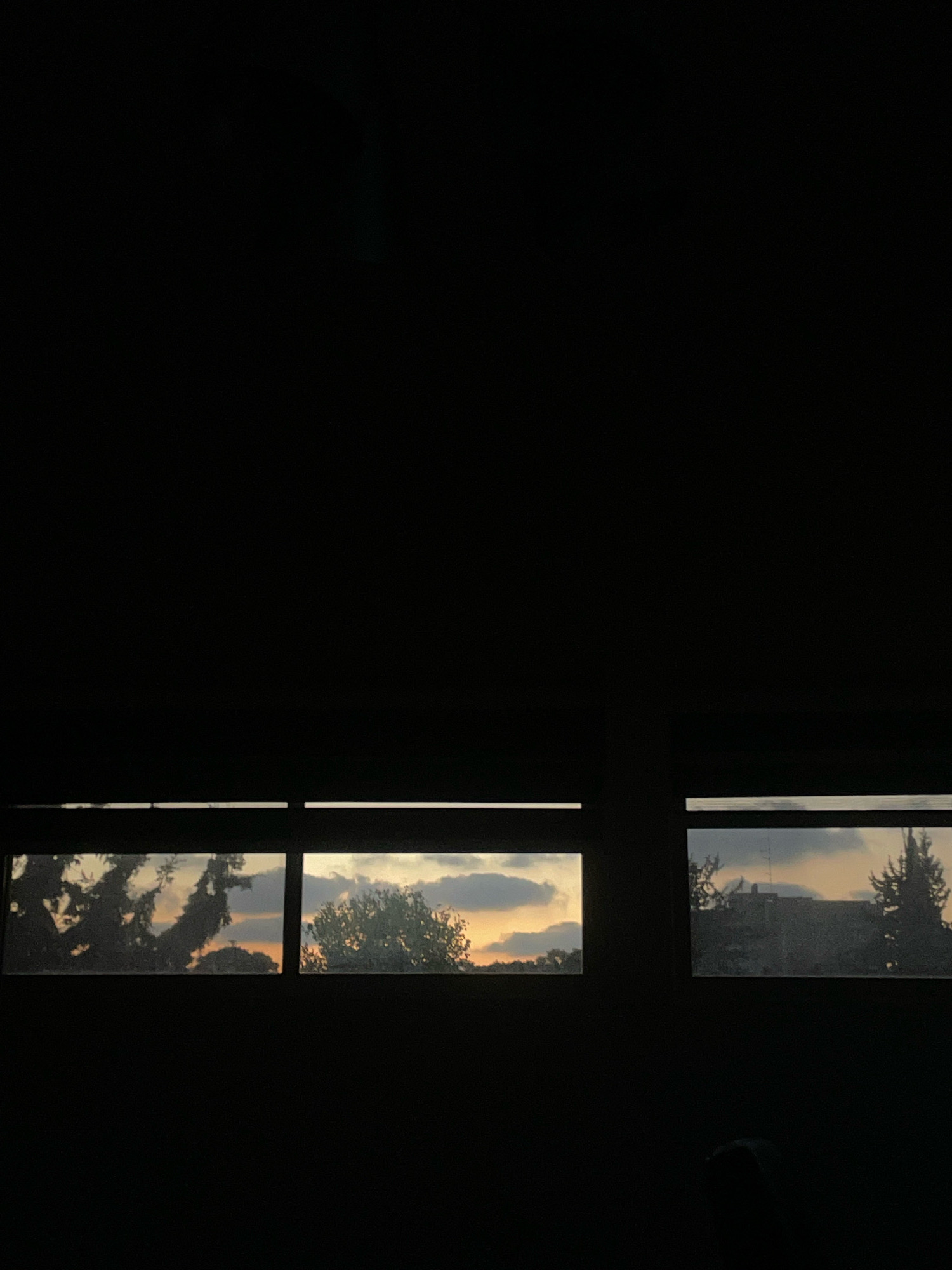 The image shows a view through a window at dusk. The sky is filled with clouds and a warm orange glow, indicating sunset. Silhouetted trees and buildings are visible against the sky. The window is divided into three sections by dark vertical bars. The left section shows a large, silhouetted tree with dark foliage. The middle section shows more trees and the sky with clouds. The right section shows a building with a few trees in front of it. The overall scene is dark and moody, with the light of the sunset providing the only significant illumination. The location is Raanana, Israel.