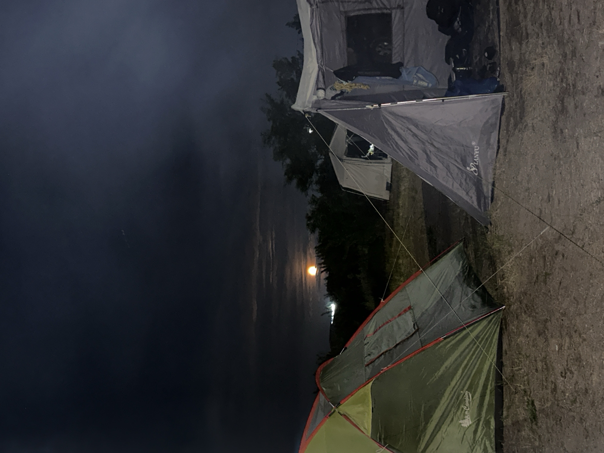 The image depicts a campsite at night, illuminated by a bright full moon visible in the dark sky. Two tents are prominently featured: a green and gray tent on the left, and a larger gray tent on the right, which appears to be open, revealing some items inside. The surrounding environment consists of dry ground with scattered vegetation. In the background, trees and distant lights are visible, suggesting a possible proximity to an urban or semi-urban area. The sky is partly cloudy, with the moon shining through. The scene is consistent with a campsite in the vicinity of the village of Kolodezny, Russia, given the natural and lightly developed landscape.