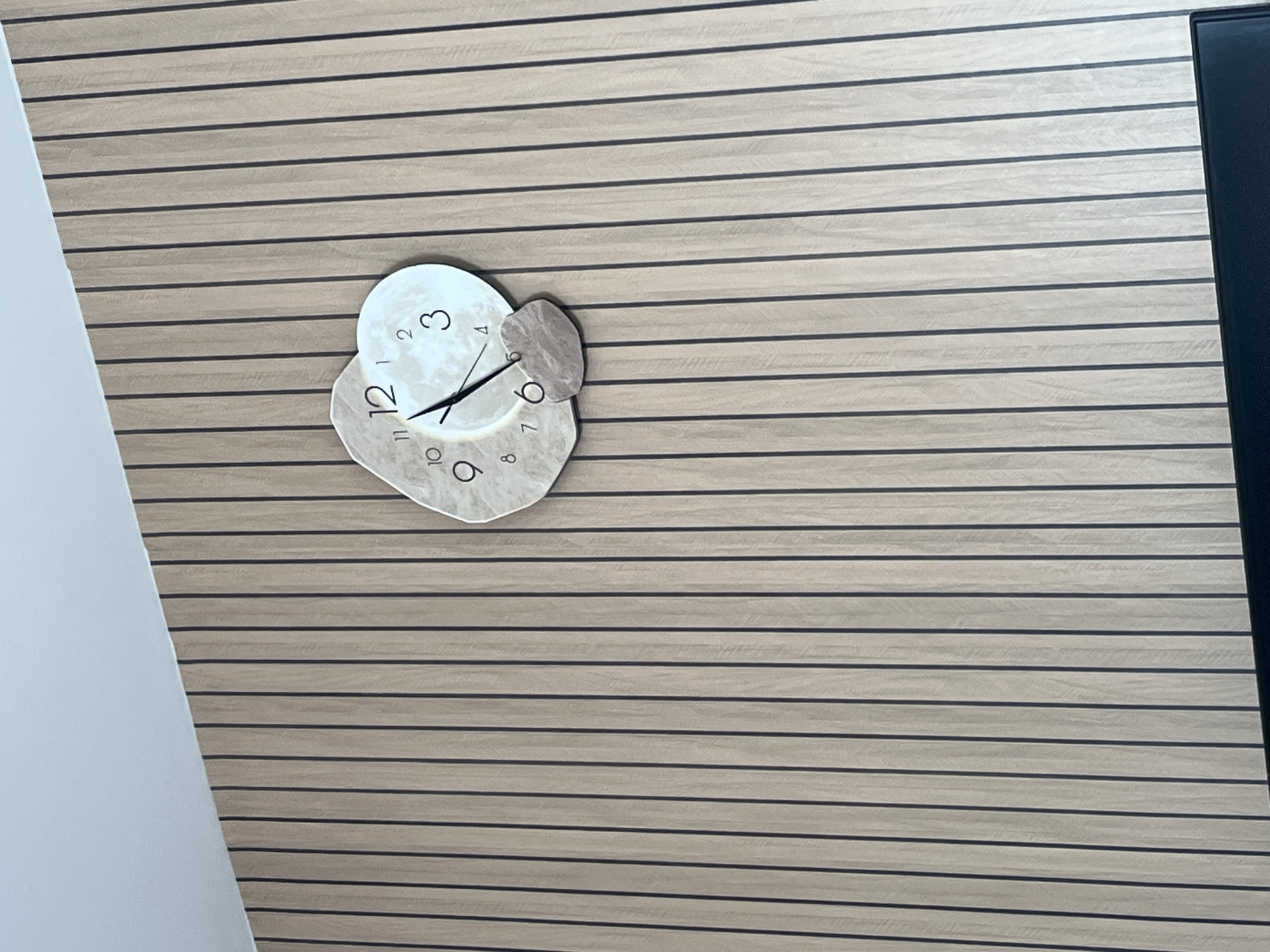 The image shows a close-up of a unique clock mounted on a vertically paneled wall. The clock has an abstract, organic shape with two distinct parts. One part is irregularly shaped and features numbers 7, 8, 9, 10, 11, and 12. The second, circular part displays numbers 2, 3, and 4, and appears to represent a moon. The clock hands are visible; the hour hand is pointing towards the number 6, and the minute hand is pointing just past the number 12. A small, pebble-like decorative element is attached to the clock near the number 6. The wall behind the clock is made of light-colored wooden panels with darker vertical lines separating them. The top of the image shows a portion of a light-colored ceiling. There are no people or any indication of the specific location within Garmisch-Partenkirchen, Germany.