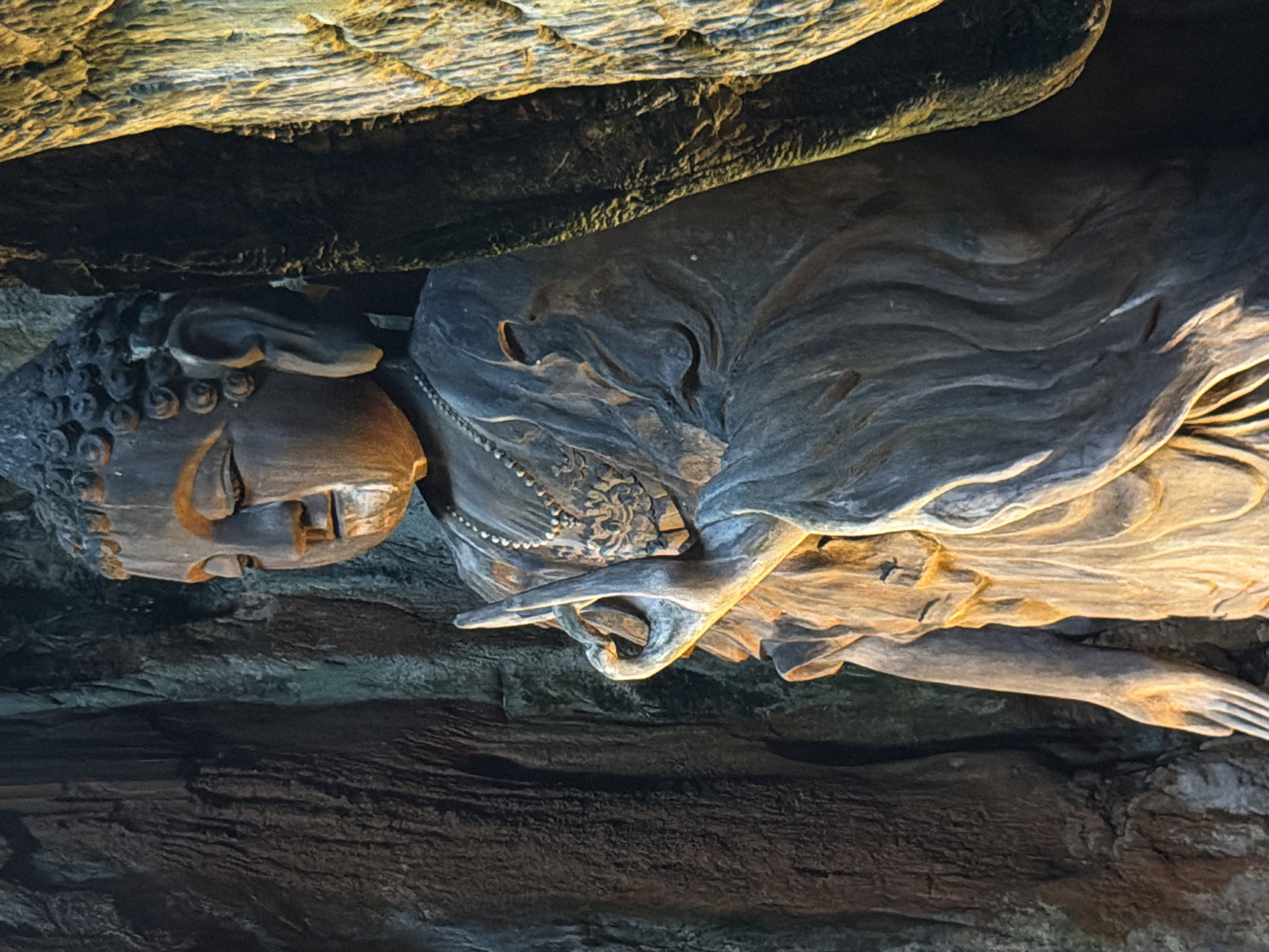 A horizontal medium shot captures a carved reclining Buddha statue, possibly made of dark wood, in the city of Đà Nẵng, Vietnam. The statue rests within a cave-like structure, with rough rock formations visible above and below. The Buddha's face is in profile, with detailed curly hair. The figure wears a necklace and robes carved with intricate designs. The right hand is raised in a symbolic gesture. The lighting appears to be artificial, highlighting the statue's texture and the cave's rough surfaces.
