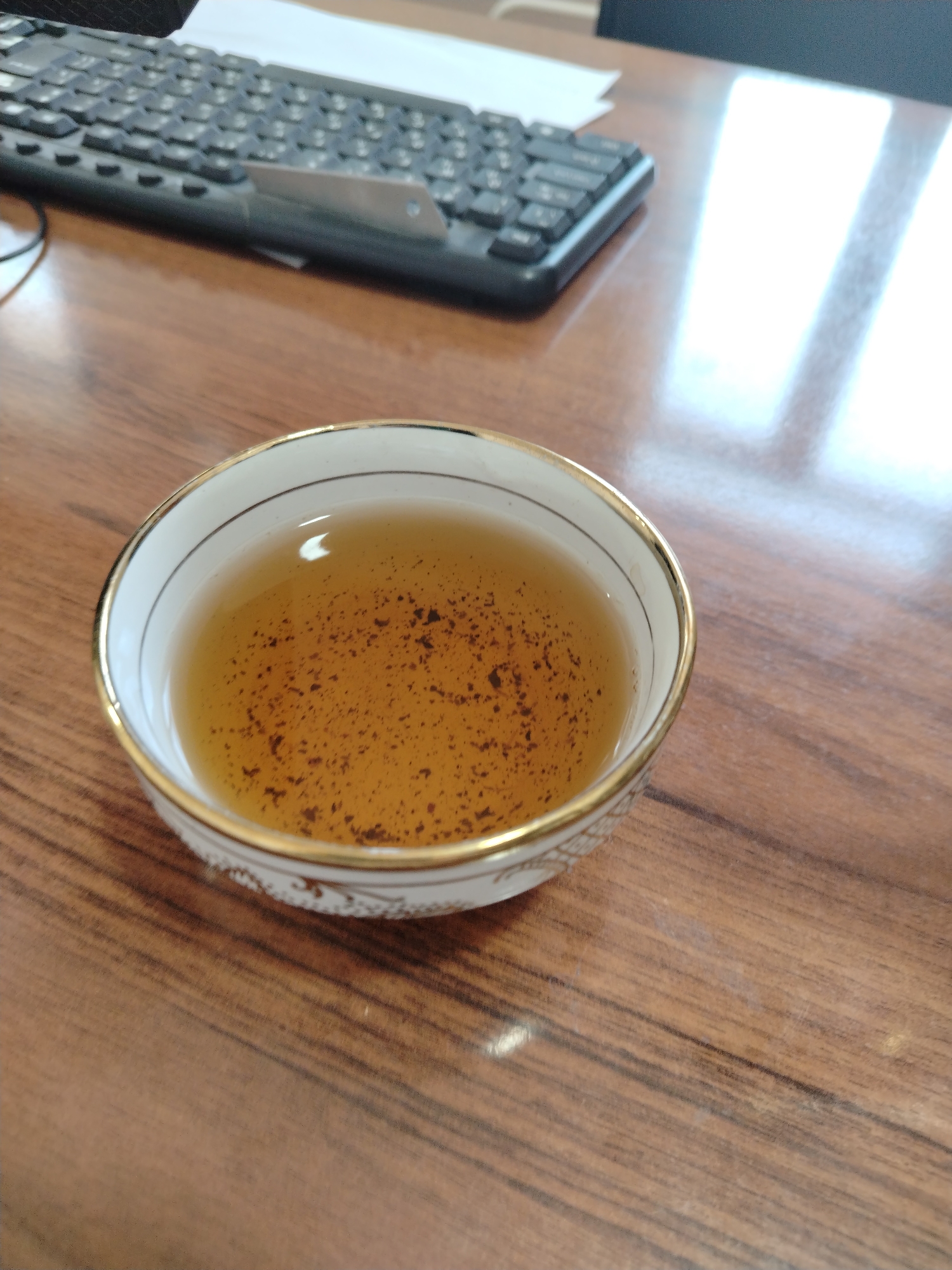 In the foreground, a white teacup with a gold rim contains amber-colored liquid with tea leaves. The cup sits on a wooden desk. In the background, a black keyboard is visible. The scene is illuminated by natural light, casting reflections on the desk. The image appears to be taken indoors, possibly in an office setting in Nurata, Uzbekistan, though no definitive identifying markers are present.