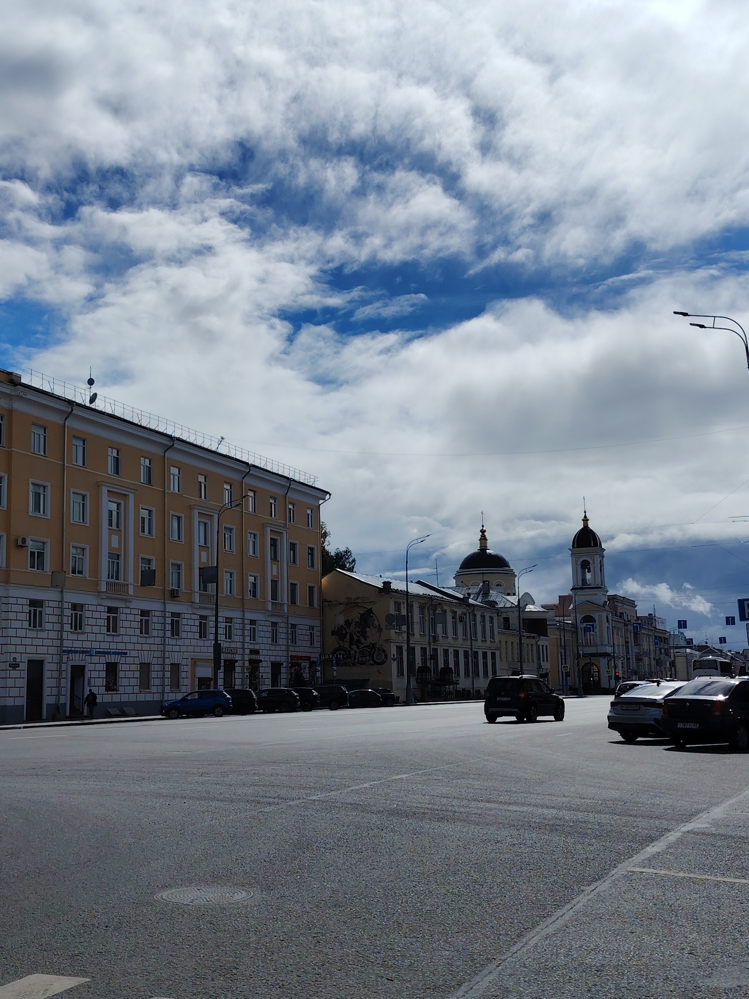 The image depicts a street scene in Tver, Russia. The foreground is dominated by a wide asphalt road with faint white lane markings. A circular manhole cover is visible on the road. To the left, a multi-story building with a yellow facade and white decorative elements lines the street. Several windows, some with balconies, punctuate the building's facade. A few cars are parked along the sidewalk in front of this building. Further down the street, a prominent church with a dark dome and a bell tower is visible. Various other buildings with different architectural styles are also present, some with visible signage. The sky above is filled with dramatic, puffy white clouds against a bright blue background. Several streetlights are scattered throughout the scene. A few individuals are visible walking on the sidewalk in the distance. The overall impression is of a moderately busy urban environment under a dynamic sky.