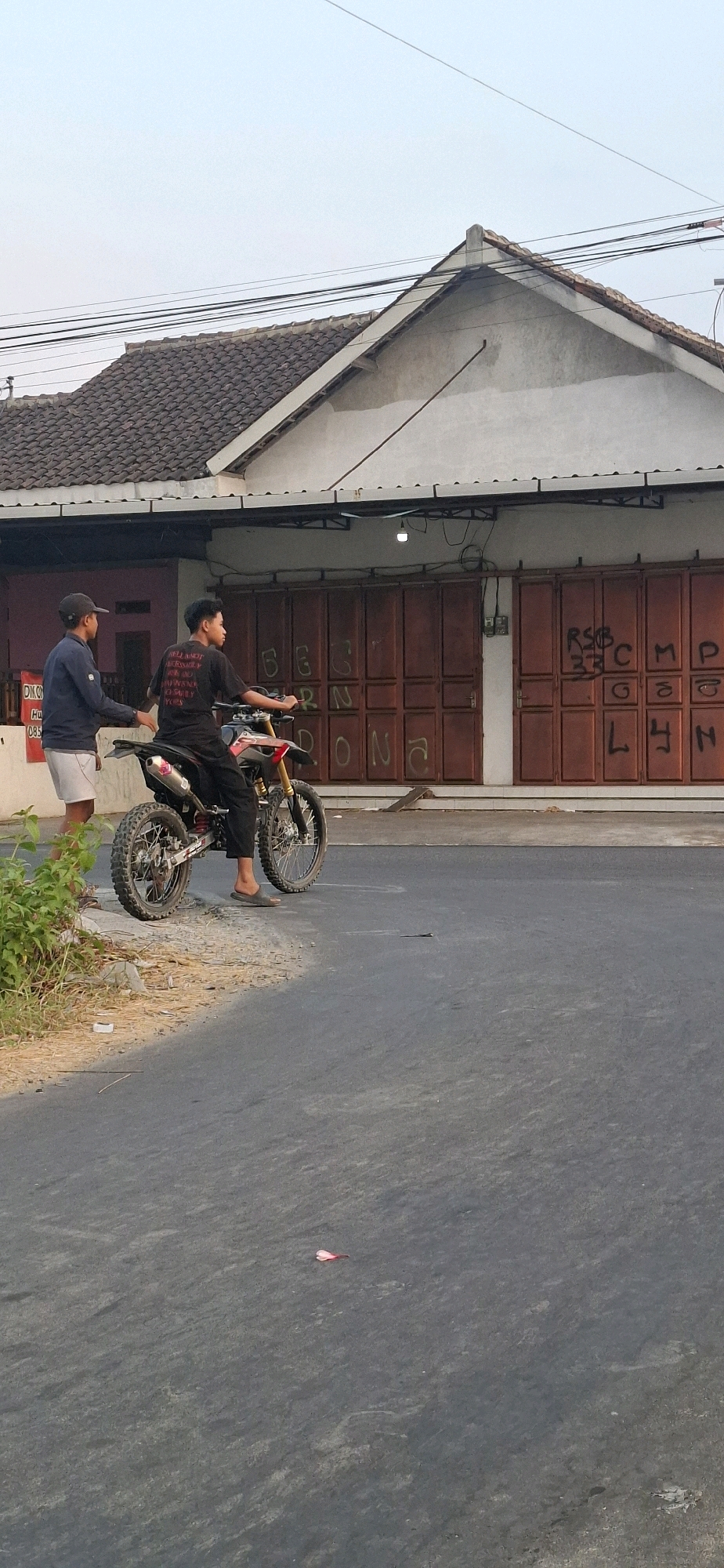 In Desa Dibal, Indonesia, on a paved road adjacent to a building with a brown metal roll-up door that is spray-painted with red graffiti, two young males are present. One male, seated on a dirt bike, wears a black t-shirt with white lettering and black pants. The other male, standing to the left, is dressed in a dark blue long-sleeved shirt and white shorts, with a black cap. He has his hands on the back of the dirt bike. The dirt bike has knobby tires and a silver exhaust pipe. Green foliage is visible on the left side of the frame. The sky is a light, overcast blue.