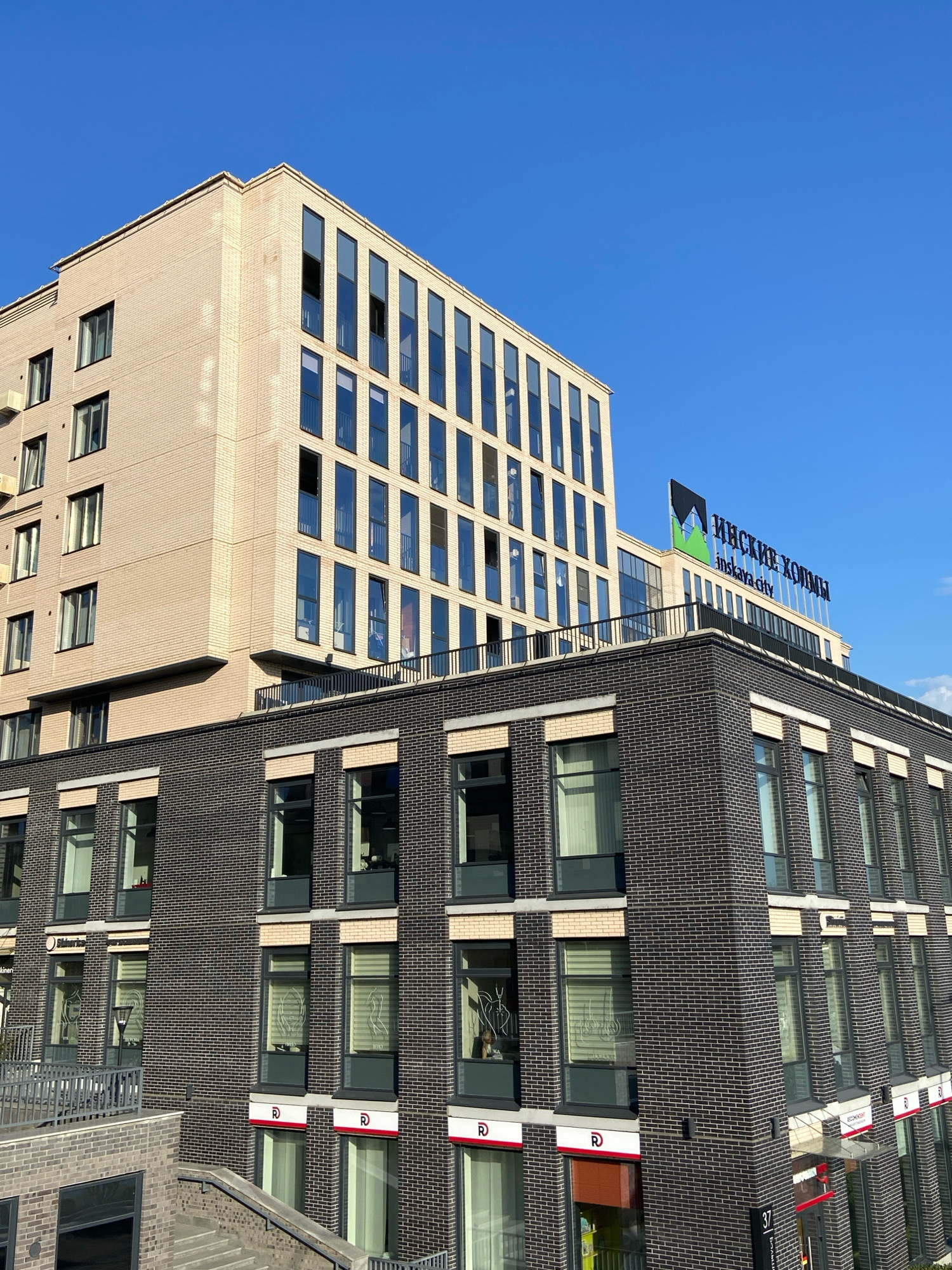 The image shows a modern, multi-story brick building under a clear blue sky. The building features a light-colored brick facade on its upper section and a darker brick facade on its lower section. Both sections have numerous large windows with dark frames. The lower section has a row of ground-floor storefronts, some with red signage and the letter "R" displayed prominently. A sign on the upper section of the building reads "ИНСКИЕ ХОЛМЫ" and "inskaya city" in English. There are no people visible in the image. The building is located in the Oktyabrsky District of Novosibirsk, Russia, specifically in the "Inskiye Kholmy" residential complex.