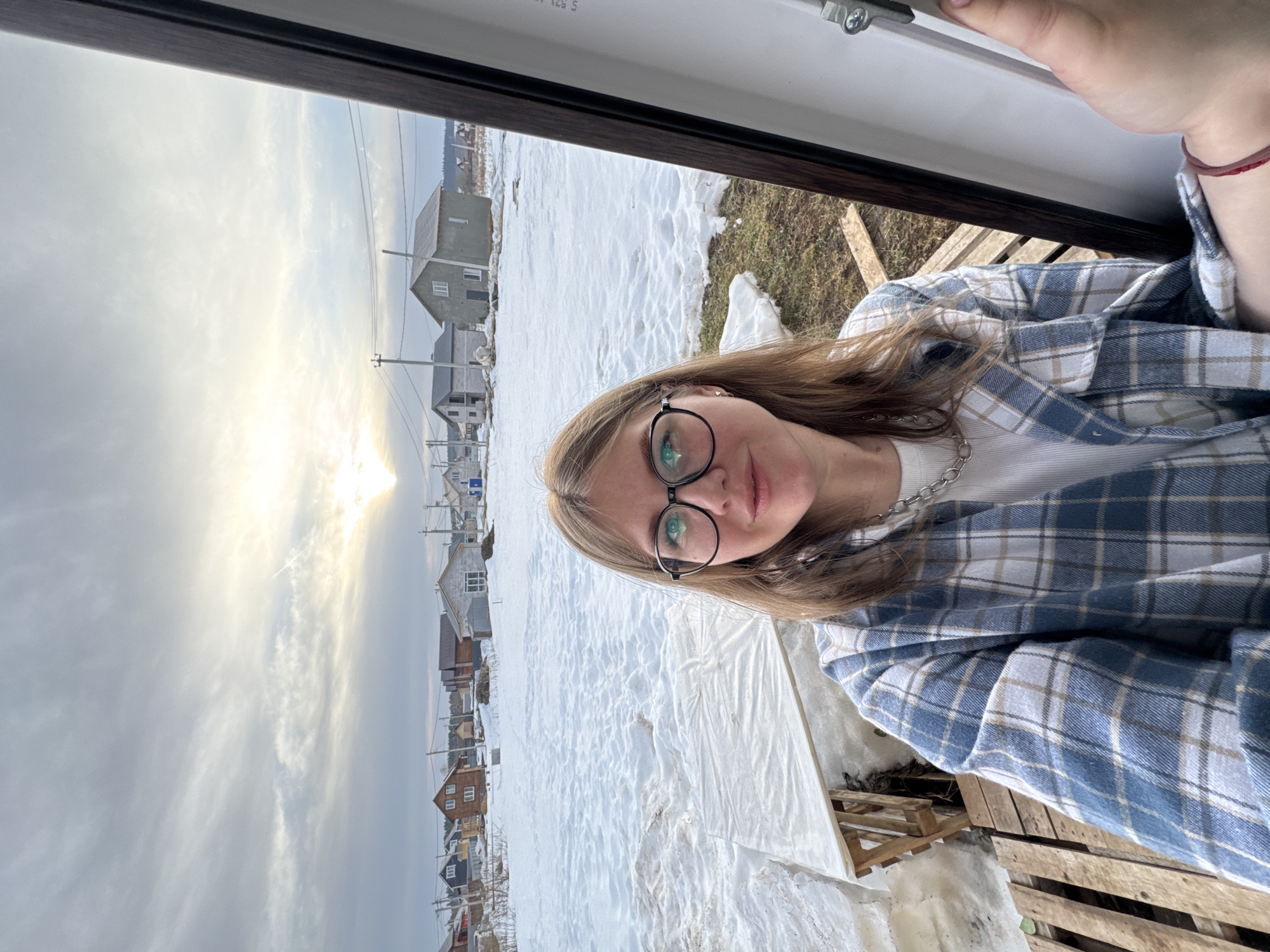 A young adult female is depicted in a close-up selfie, taken from an elevated position looking outwards from an open window or door frame in Gornyi (Dvurechenskoe S/P), Russia. She has light brown hair, wears black round-frame eyeglasses, a white ribbed shirt, and an unbuttoned blue, white, and tan plaid shirt. A silver-colored chain necklace is visible, and a red string bracelet is on her right wrist, which is partially visible holding the dark brown and white window/door frame.
The exterior scene reveals a winter or early spring landscape. The ground immediately outside the opening features melting snow, exposed brown earth, and wooden construction pallets. Further back, a large, flat, rectangular object covered by white plastic sheeting rests on supports within a snowy area. The mid-ground and background consist of a residential area under a partially overcast sky with the sun visible on the right, casting a bright glare. Numerous houses, some complete and some under construction (concrete block, wooden, and sided structures), are scattered across a snow-covered terrain. Utility poles and power lines are present throughout the housing area. A distant line of dark trees marks the horizon.