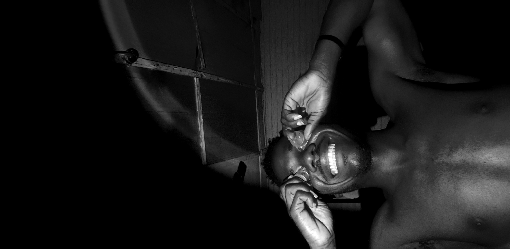 This is a black and white, horizontal, close-up photograph of a Black man's face and upper chest. The image is oriented such that the man's head is towards the bottom right of the frame, and his body extends upwards and to the left. His eyes are wide open, and he is smiling broadly, revealing his teeth. His mouth is slightly distorted as his fingers are pulling at the corners of his lips. He has a short beard and a dark band on his upper left arm.
The background is dark, with a portion of what appears to be a window frame or a doorway visible in the upper left quadrant. There are streaks of light coming through this structure, casting shadows. The texture of the man's skin is visible, with highlights and shadows emphasizing his features. The overall mood of the image is intense and somewhat unsettling due to the extreme close-up and the man's expression and action. The lighting suggests an indoor setting, possibly at night or in a dimly lit room. There is no visible text in the image.