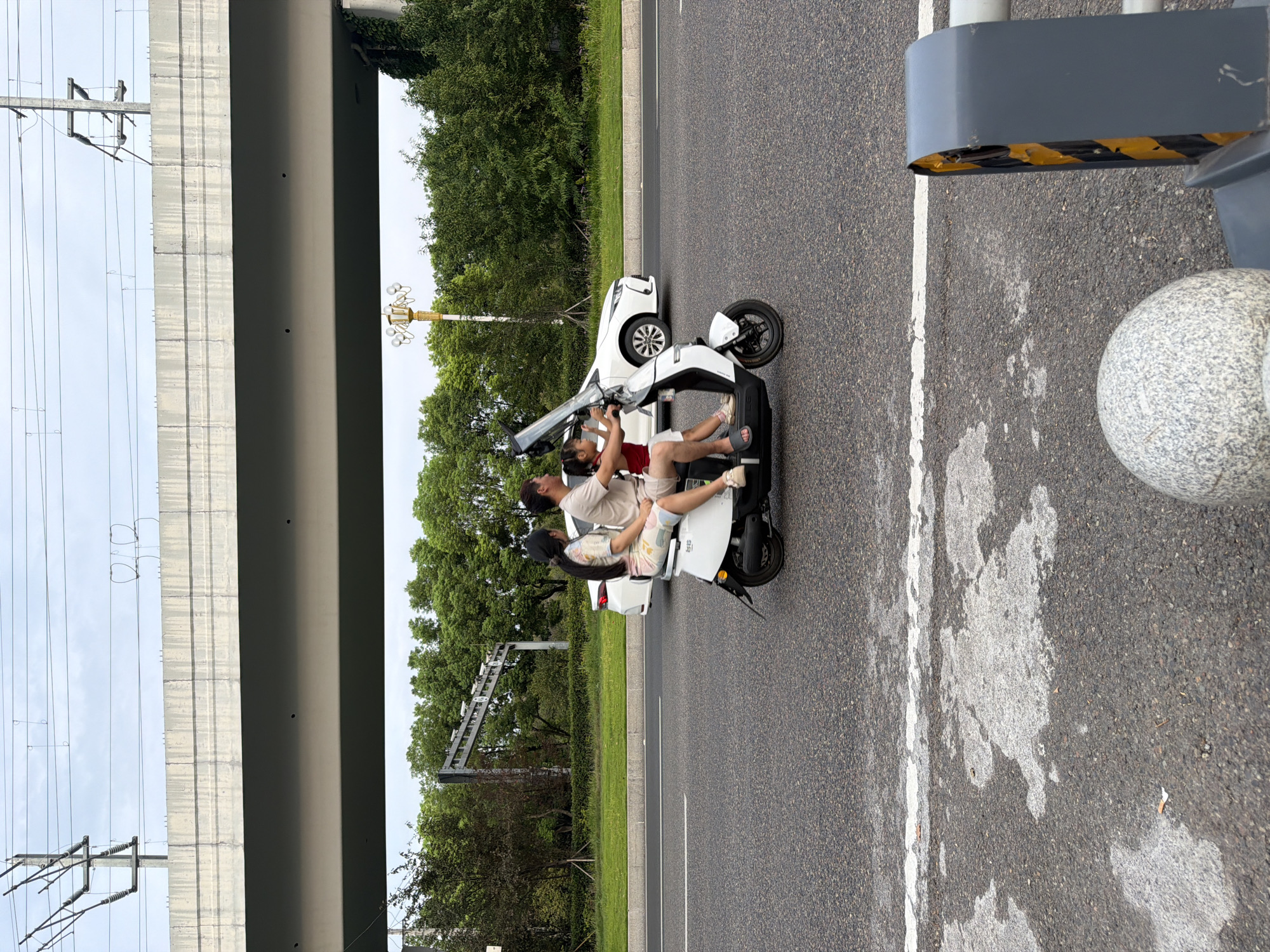 In Jiangjin, China, on a multi-lane road with a white lane marker, a family of three is riding a white electric scooter under a concrete overpass. The father, wearing a light-colored t-shirt and shorts, is driving the scooter. Next to him, a woman in a patterned dress is seated behind him. In front of the father, a young girl in a red top and white shorts is also seated on the scooter, holding onto the handlebars. A white car is visible driving in the lane next to them. Overhead, power lines crisscross the sky, supported by utility poles. The environment is lush with green trees lining the road, and the sky is overcast. In the foreground, a grey bollard and part of a metal barrier are visible on the right side of the frame.