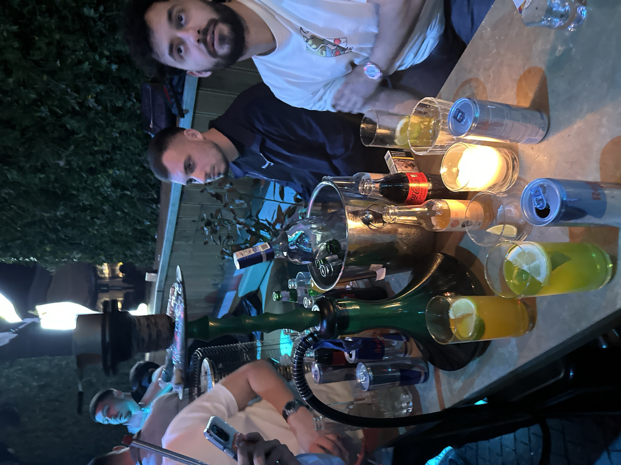 Two men are seated at a table in an outdoor setting in Plovdiv, Bulgaria. The table is cluttered with various items, including a hookah, several glasses with drinks, cans of Red Bull, bottles of Coca-Cola and other beverages, a candle, and an ice bucket filled with bottles. One man is looking at the camera with a beard and a white shirt and is on the right side of the image. Another man is seated and looking at the table. Another man is seated at the table and is holding a cell phone.