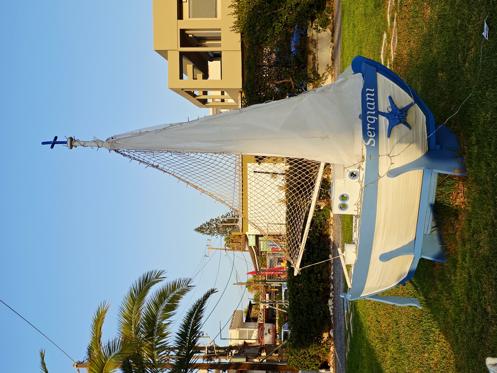 In Malia Municipal Unit, Greece, a decorative model sailboat is positioned on a grassy area, likely in a public space or garden. The sailboat is white with a blue hull and is mounted on blue stands. A white sail is unfurled, and a blue starfish decoration is affixed to the bow, which is inscribed with the name "Sergiani." A blue cross tops the mast. Behind the sailboat, a low green hedge lines a pathway and a wall. Further in the background, buildings and other structures are visible, including a modern building with a beige facade and dark window frames, and a smaller building with an "ATM" sign. Palm trees and utility poles with numerous wires are also present in the scene. The lighting suggests it is late afternoon or early evening, with the sun casting long shadows. There are no people or animals depicted in the image.