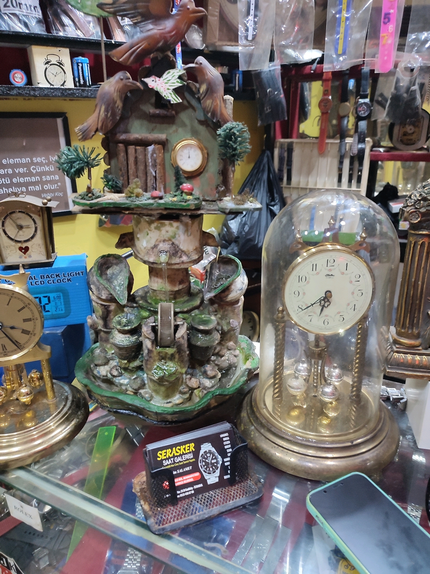 A detailed, forensic, matter-of-fact description of the scene is as follows:
The image captures a close-up, eye-level view of a display of antique clocks and decorative items, likely within a shop in Kadıköy, Turkey. The foreground is dominated by a glass countertop reflecting various items.
On the left, a golden pendulum clock with a white face and Roman numerals is partially visible. To its right, a blue box with "BACK-LIGHT LCD CLOCK" printed on it is seen, displaying a digital time of "02:21". Further right, a multi-tiered, green and brown ceramic fountain-like structure is the central focus. This structure is adorned with small ceramic birds perched on top and appears to be functional, with water flowing down. Various small potted plants and mushrooms are incorporated into its design.
To the right of the fountain, a large, ornate clock encased in a glass dome is prominent. This clock has a white face with black Arabic numerals and floral detailing, and a visible pendulum mechanism below. Behind this clock, other watches and decorative items are visible, including what appears to be a tall, fluted decorative column.
Scattered on the glass surface are various business cards and labels. One prominent card advertises "SERASKER SAAT GALERISI", indicating a watch gallery, and lists brands like Omega, Citizen, and Seiko, along with a contact number and address in Kadıköy, Istanbul. Another small white label with "ROLEX" is also visible.
A black trash bag is visible in the background, along with shelves holding more watches and items, suggesting a cluttered retail environment. A smartphone with a light green case lies on the lower right of the countertop. The lighting appears to be artificial, casting a warm glow on the displayed items. No people are visible in the image.