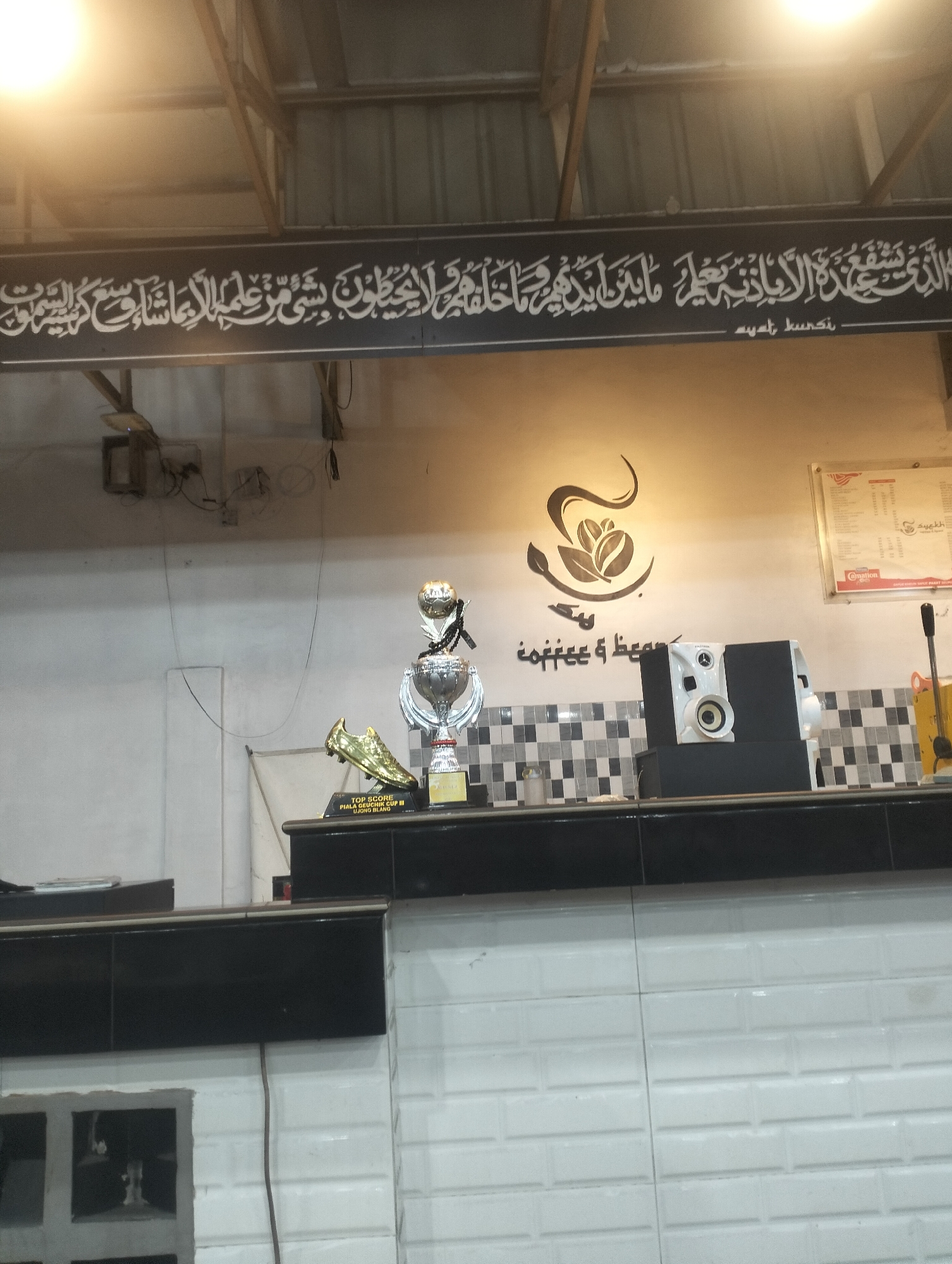 The image depicts the interior of a coffee shop in Lhokseumawe, Indonesia. Above the serving counter, a sign displays the coffee shop's logo and name in Arabic and English, "coffee & beans." A black banner with white Arabic calligraphy, presumably the Ayat al-Kursi, is hung higher up. The counter displays a trophy and a speaker. The lower part of the counter is white-tiled. Above the counter is a black and white checkered design. A menu is visible on the wall.